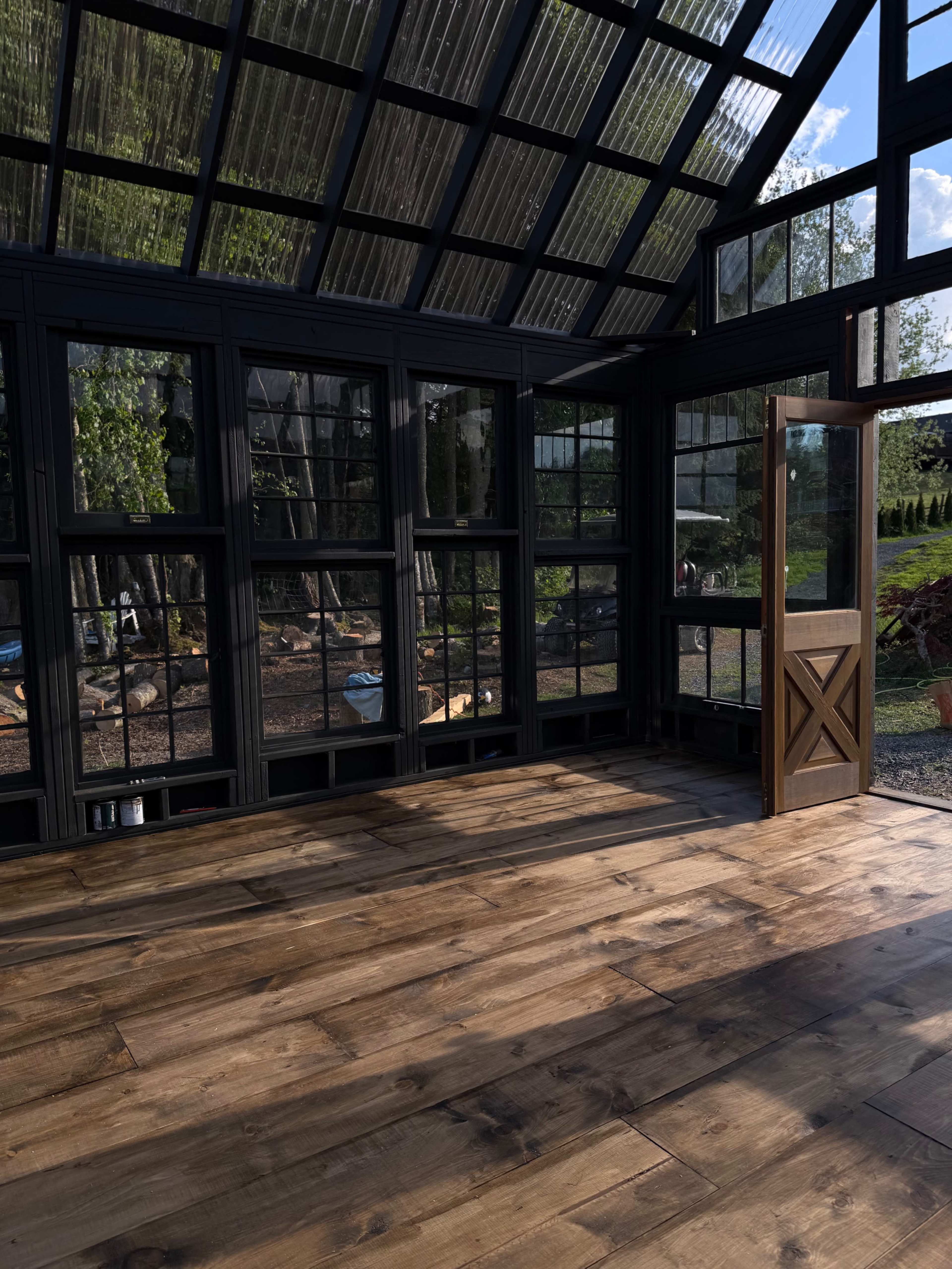 The image shows a spacious interior of a greenhouse with wooden flooring, large windows, and an open door leading outside.