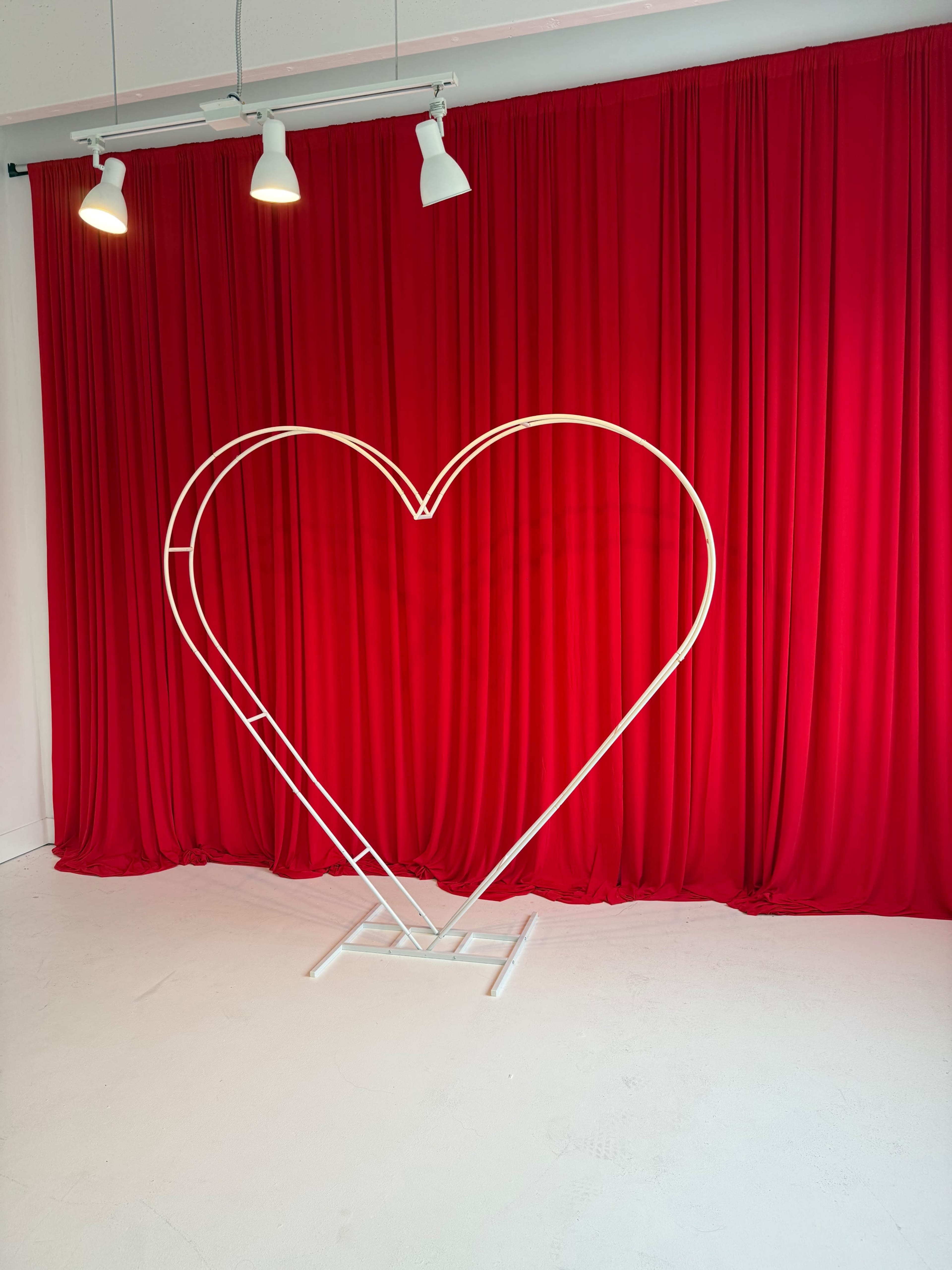 A large, white heart-shaped frame stands in front of a bright red curtain in a minimalist room.