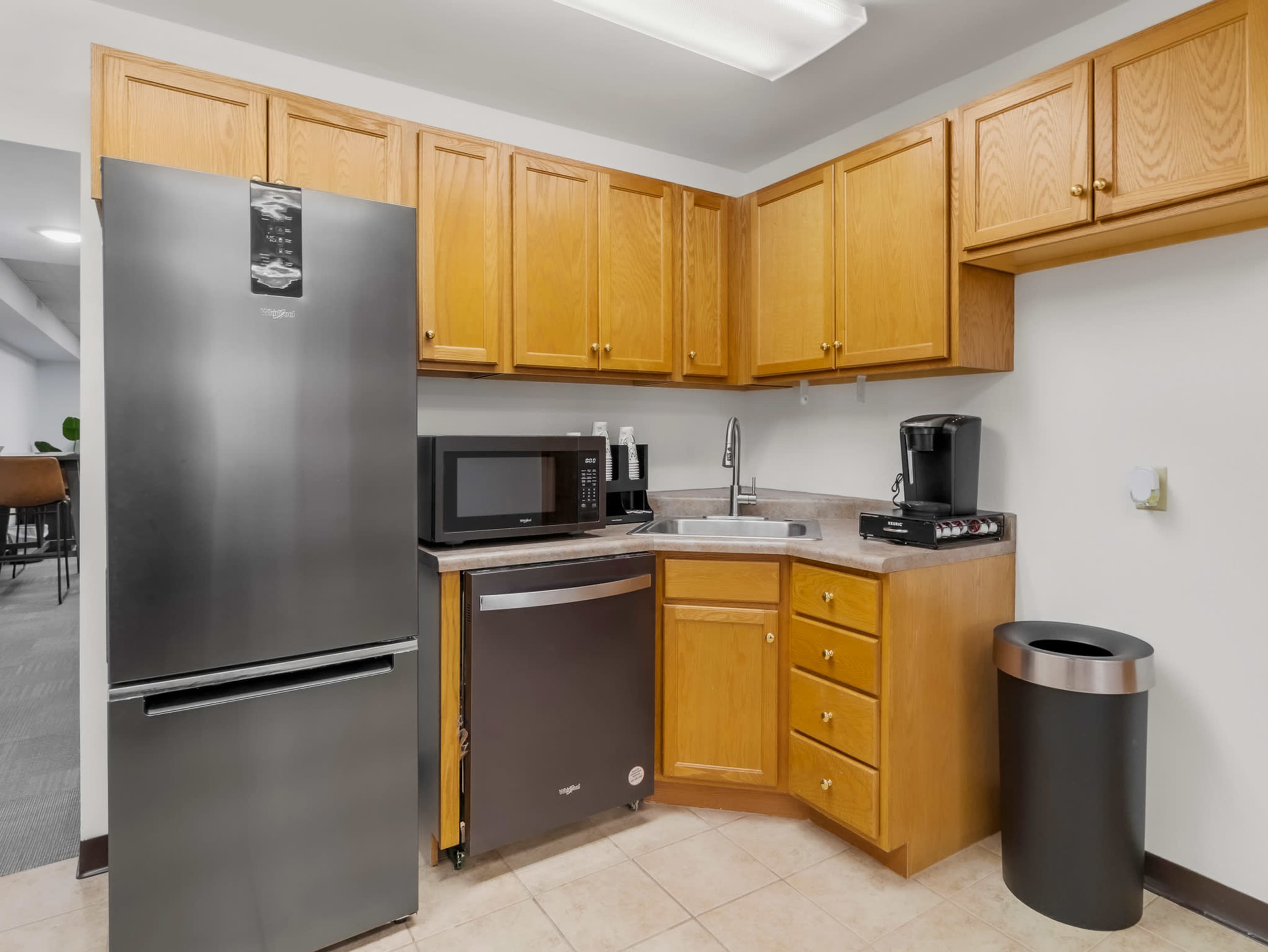 The image shows a compact kitchen featuring wooden cabinets, a black refrigerator, a microwave, a sink, and a coffee maker.