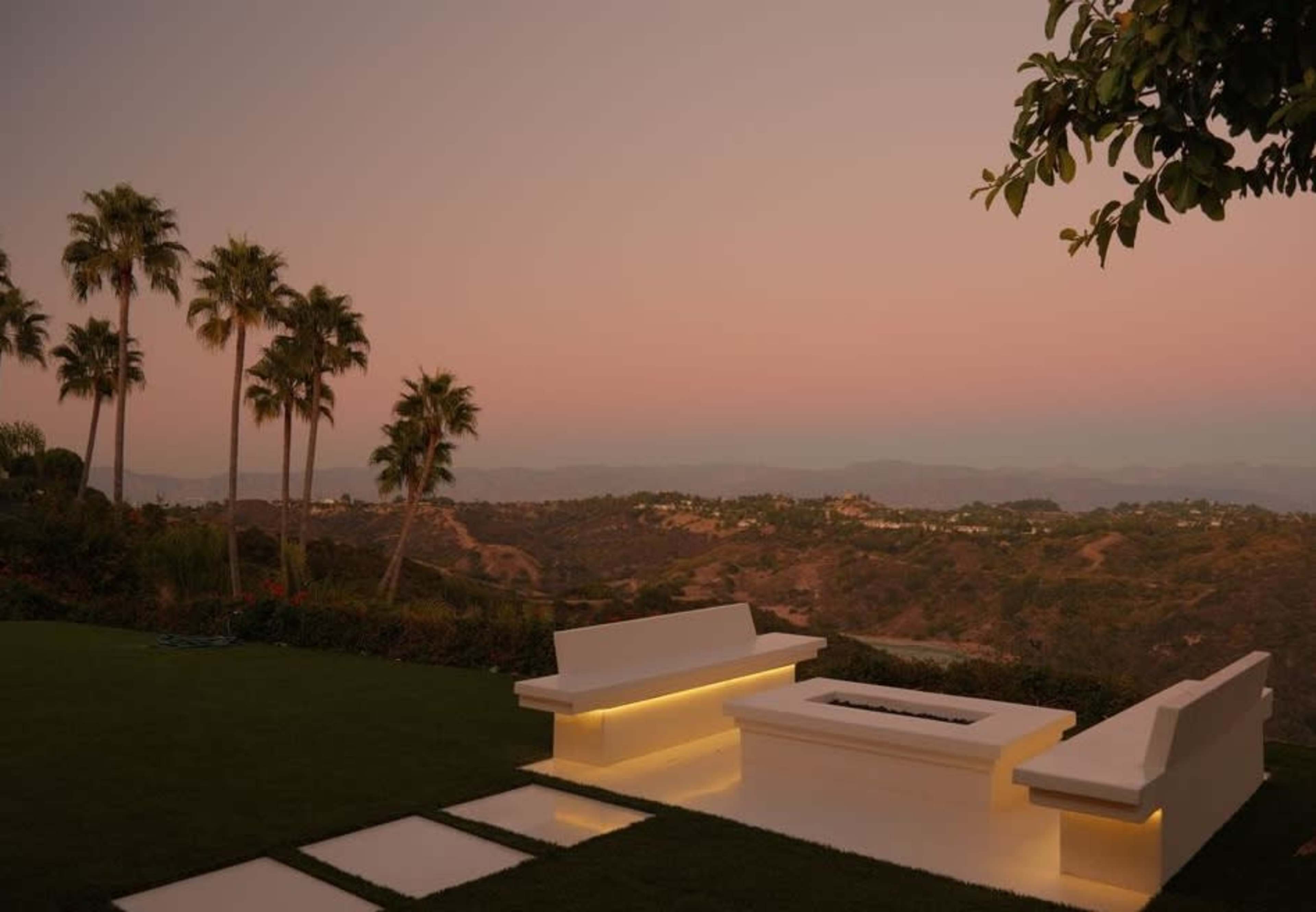 A modern outdoor seating area features white benches and a fire pit, overlooking a panoramic view of hills and palm trees at sunset.