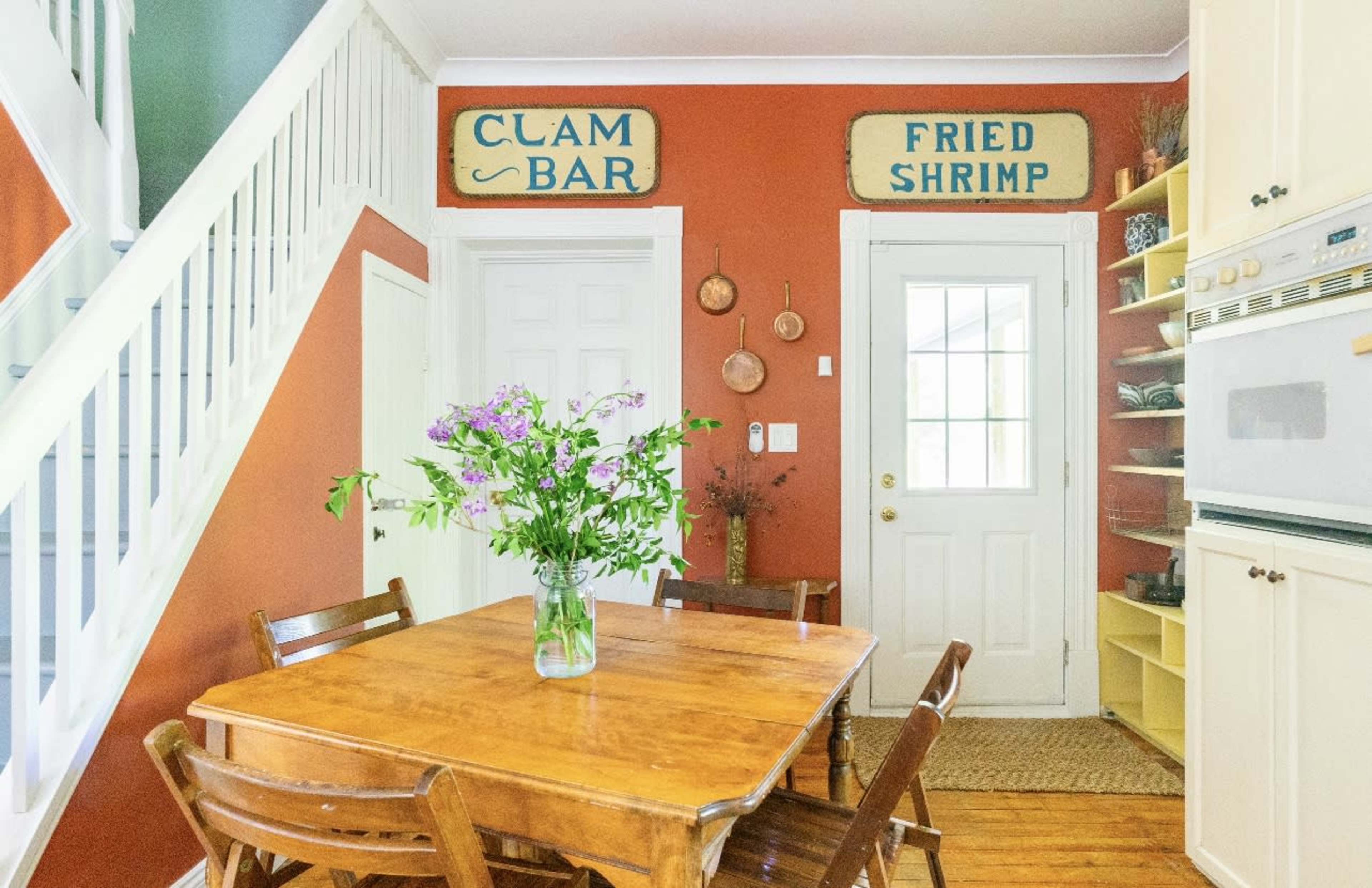 A wooden dining table is surrounded by chairs in a room with orange walls, featuring signs labeled "CLAM BAR" and "FRIED SHRIMP" above the doorway.