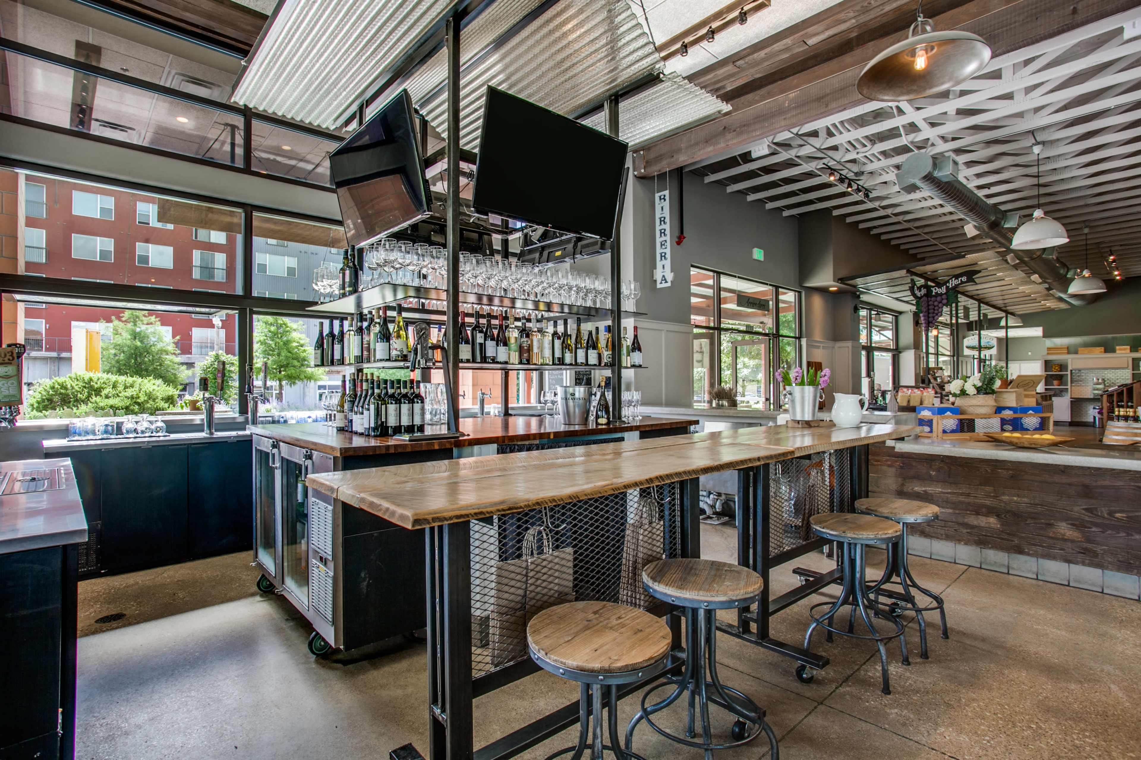 A modern restaurant bar with a wooden countertop, bar stools, and a wall of shelves filled with bottles, surrounded by large windows letting in natural light.