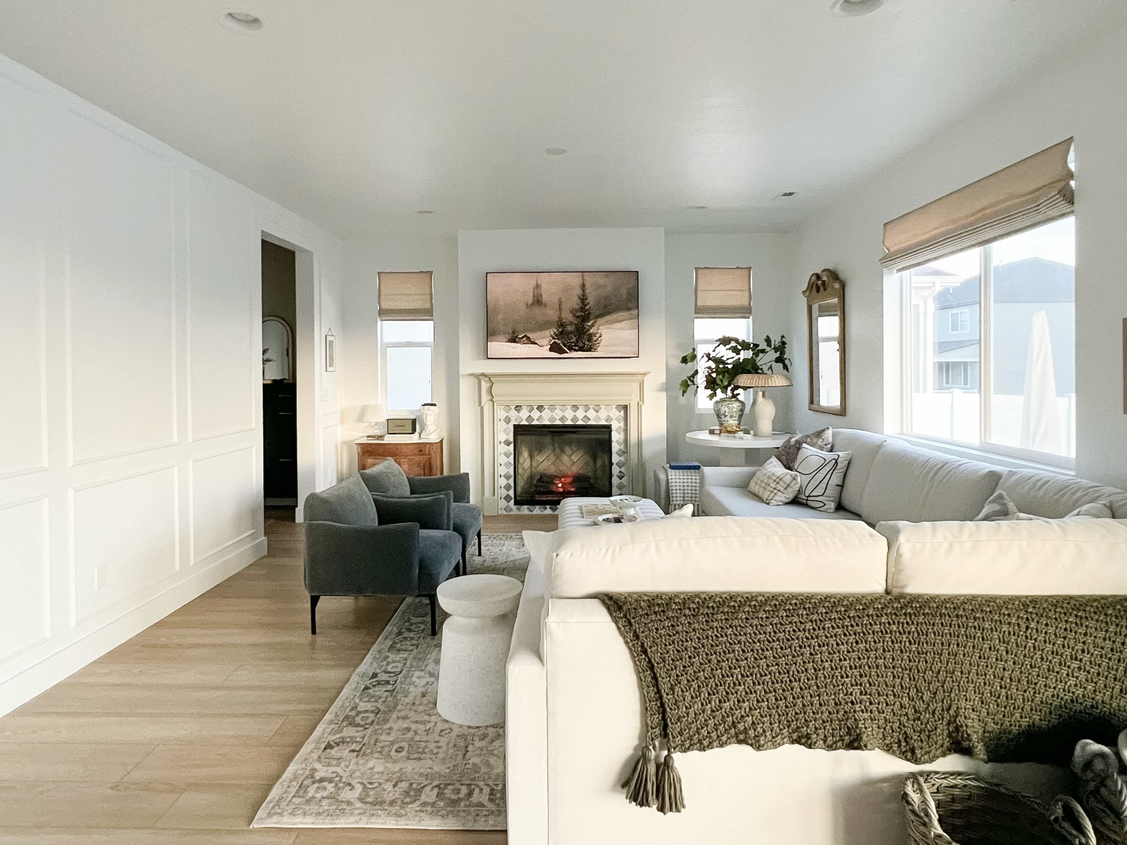 A bright living room featuring a large white sectional sofa, a fireplace with artwork above it, and two gray armchairs, all complemented by natural light from large windows.