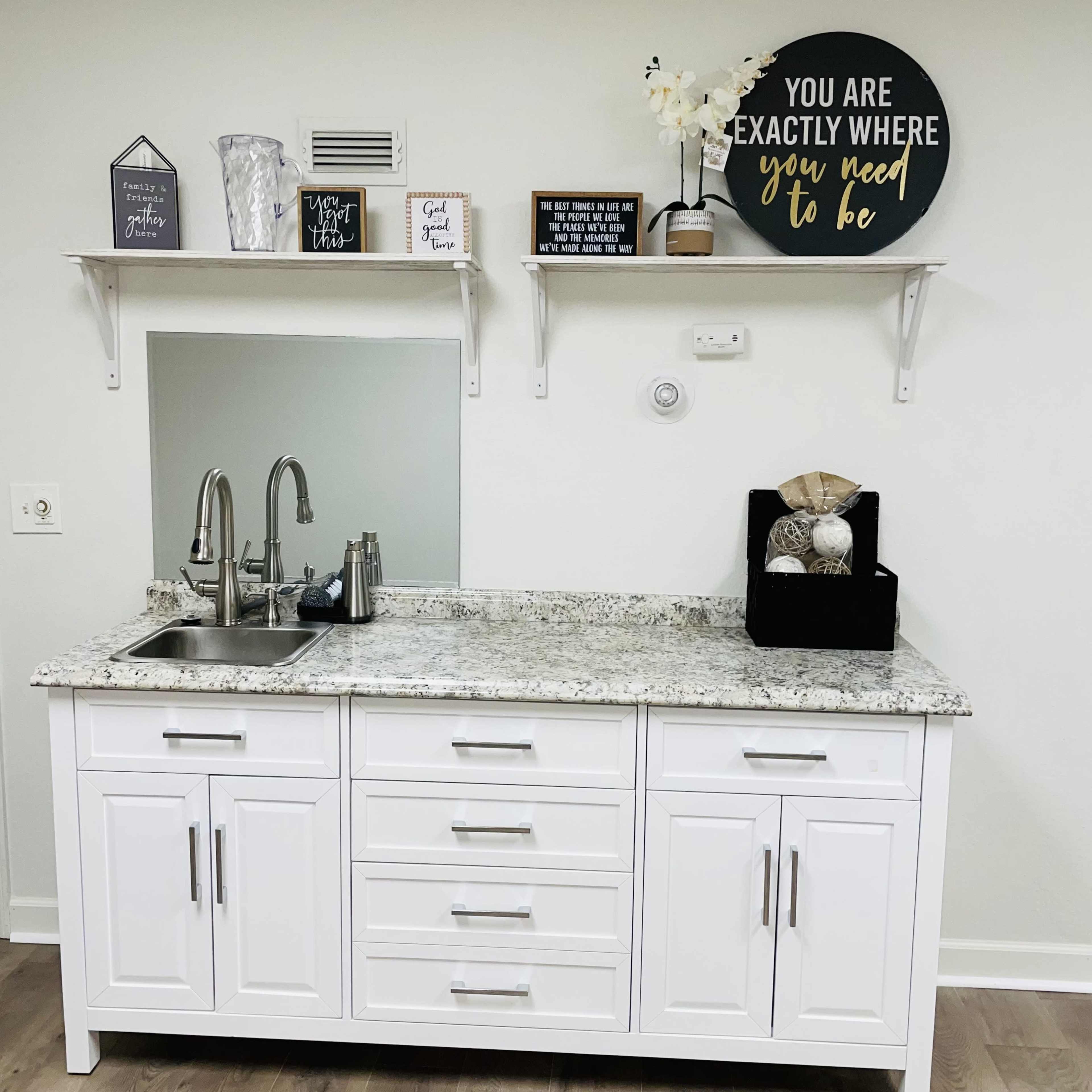 A modern kitchenette with a white cabinet, a granite countertop, a sink, and decorative wall shelves displaying various signs and a decorative basket.