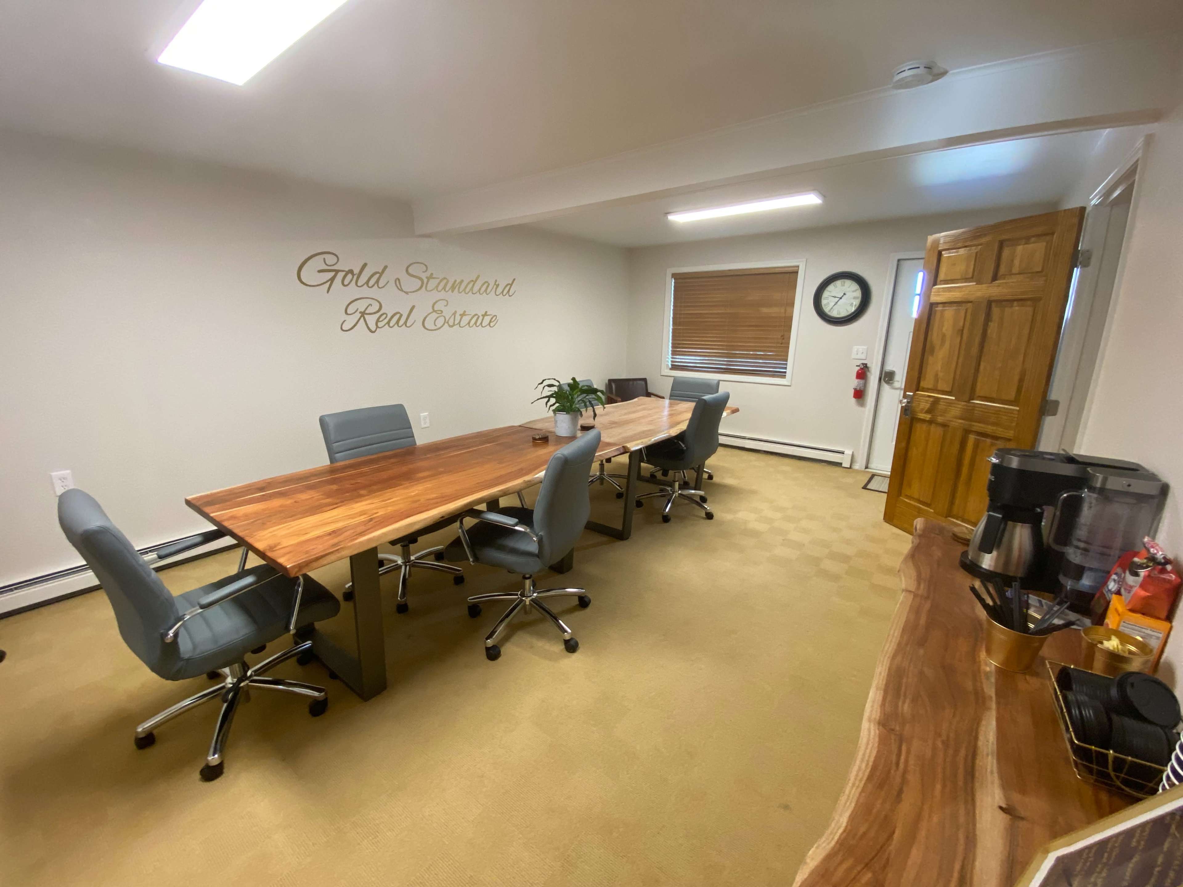 A modern conference room features two wooden tables, four office chairs, and a beverage station against a wall with the text "Gold Standard Real Estate."
