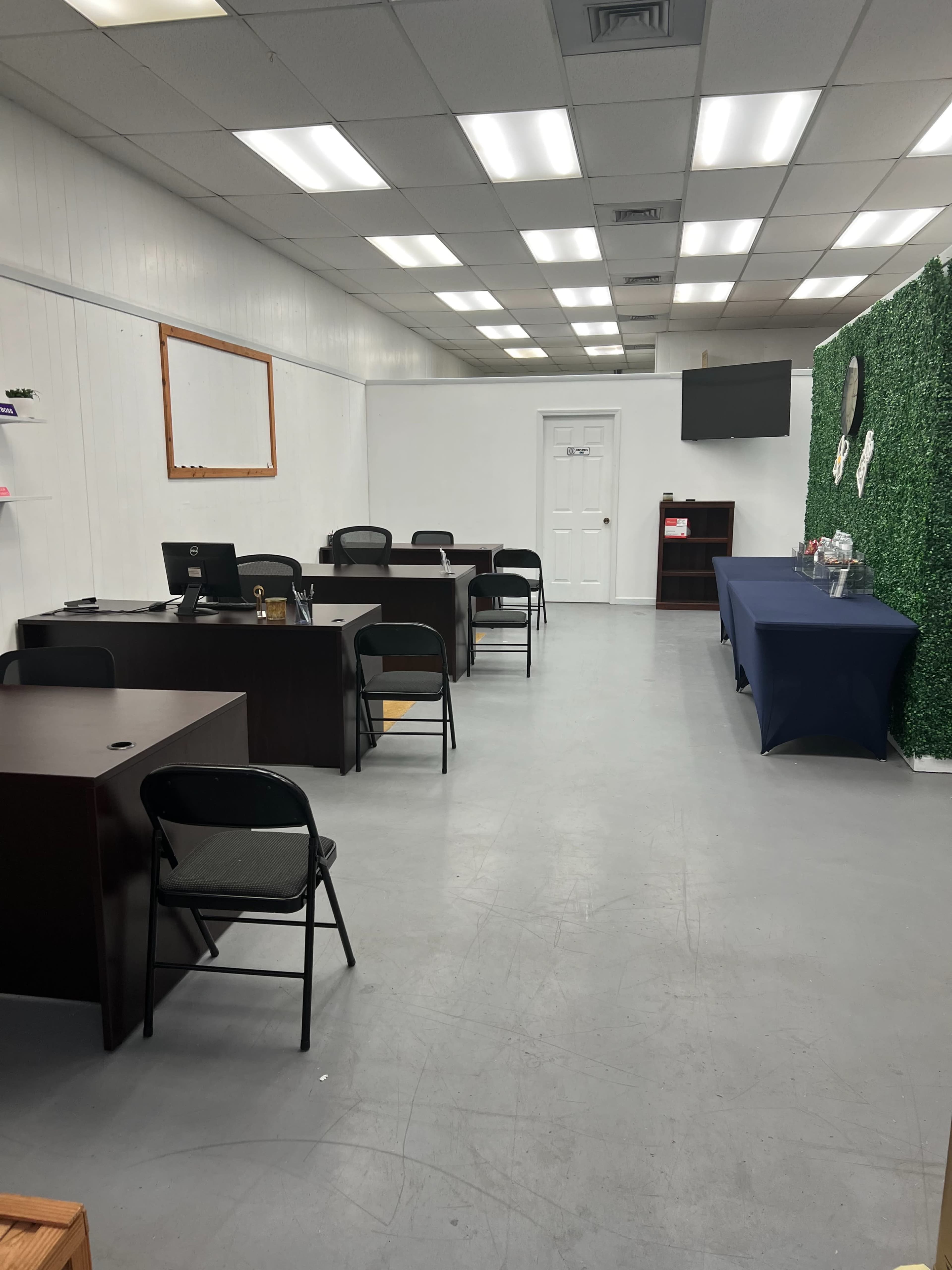 The image shows a moderately sized office space with several desks and chairs arranged in rows, a small refreshment table at the end, and a green wall feature on one side.