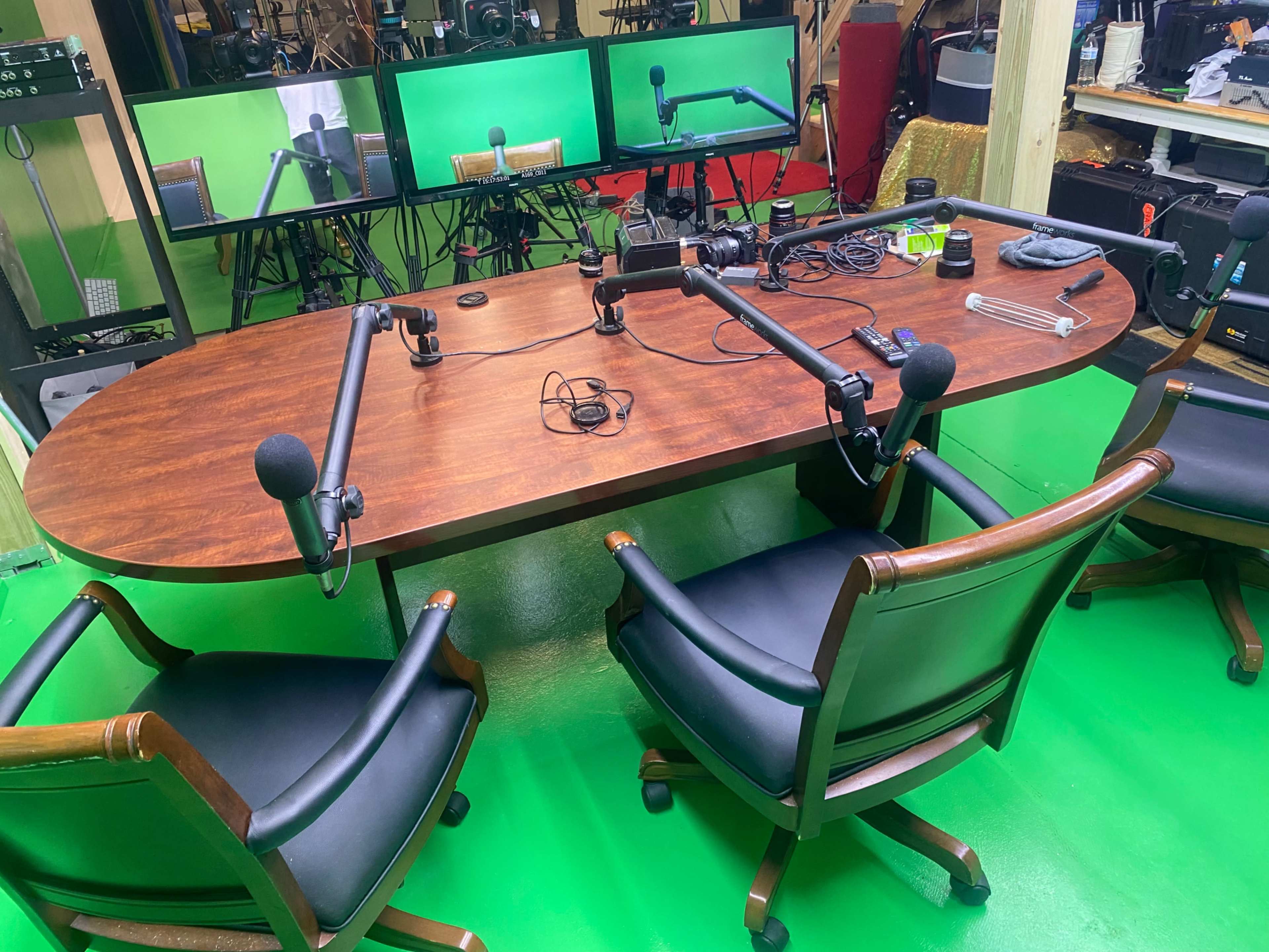 A wooden oval table with three chairs is set up in a studio, surrounded by microphones and multiple monitors displaying green screens.