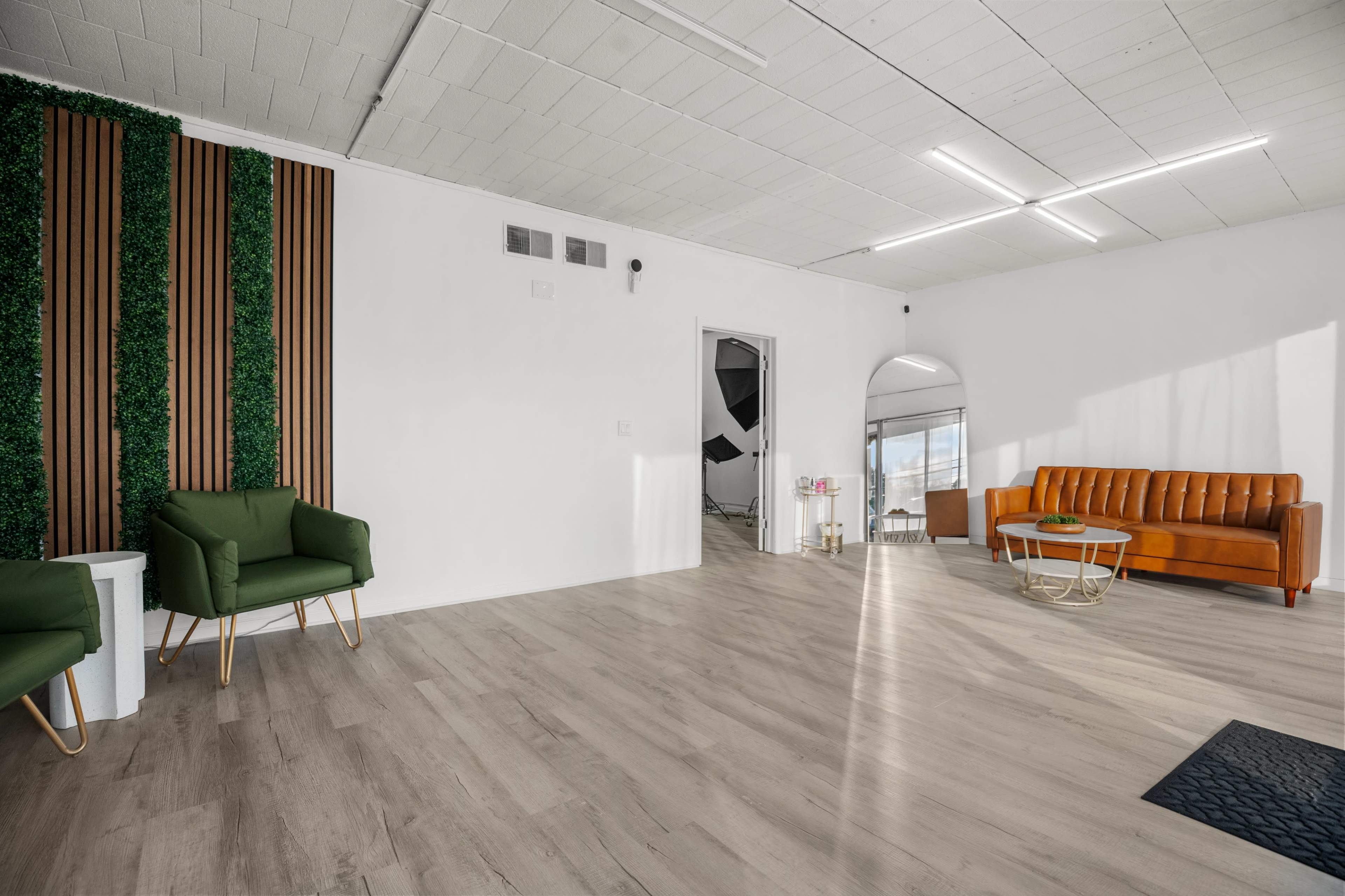 The image shows a modern, minimalistic interior space featuring a wooden floor, green accent chairs, an orange couch, and a wall adorned with vertical wooden panels and greenery.