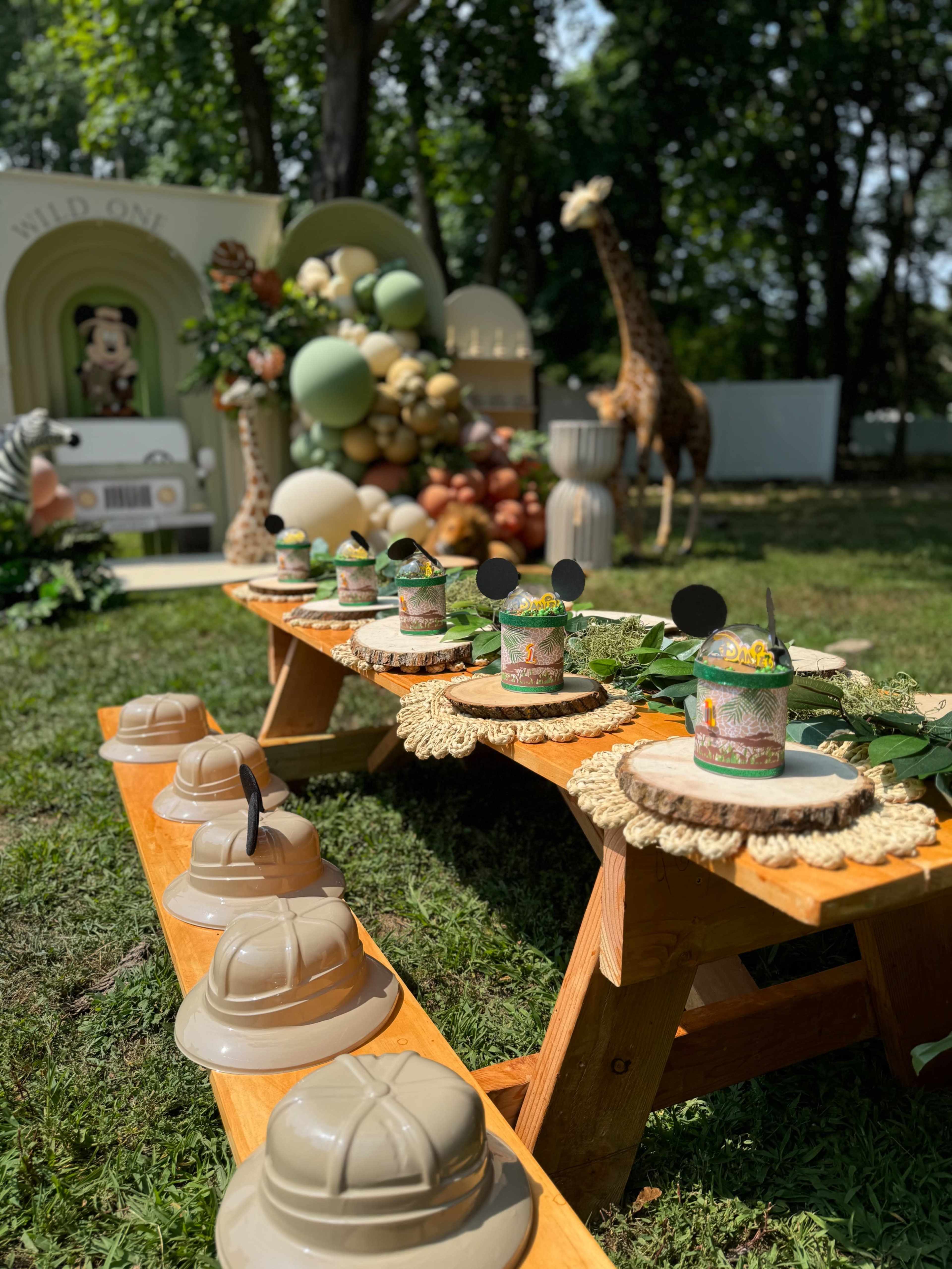 A festive outdoor table set with themed party hats and decorative cups, surrounded by a backdrop of greenery and balloon arrangements.