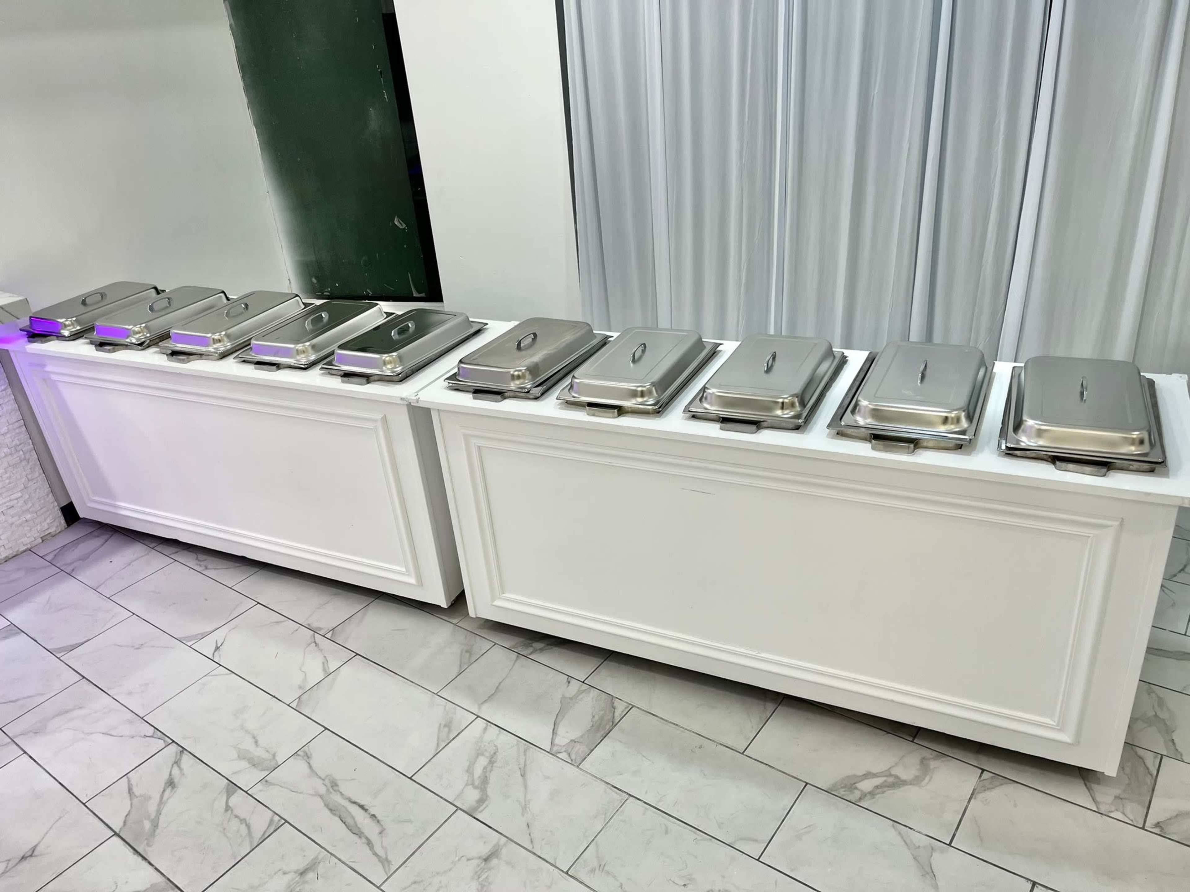 A line of covered serving trays is arranged on a white table in a dining area.