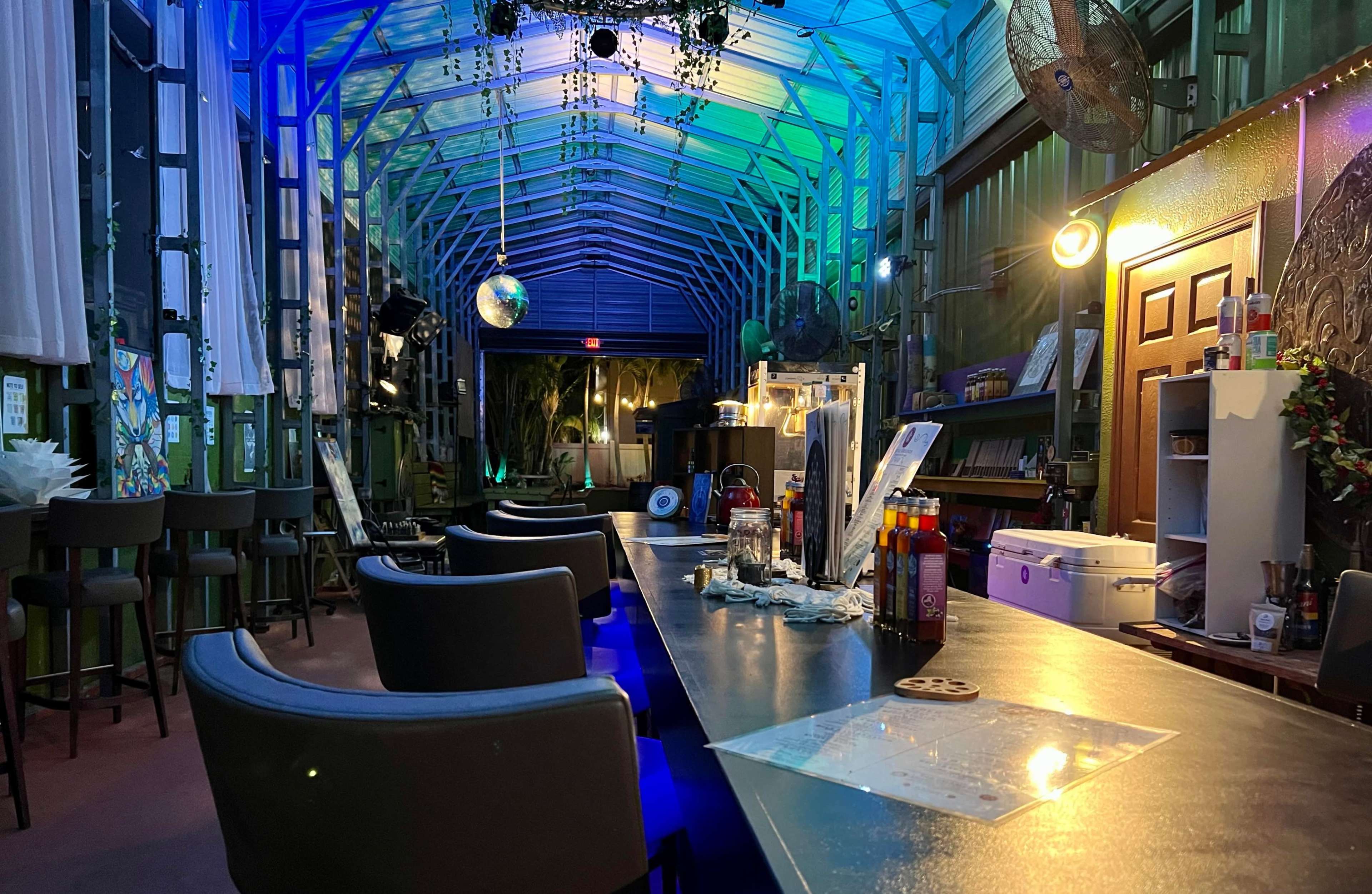 A bar area in a green, well-lit room with high ceilings, featuring a long counter, stools, and decorative lights.