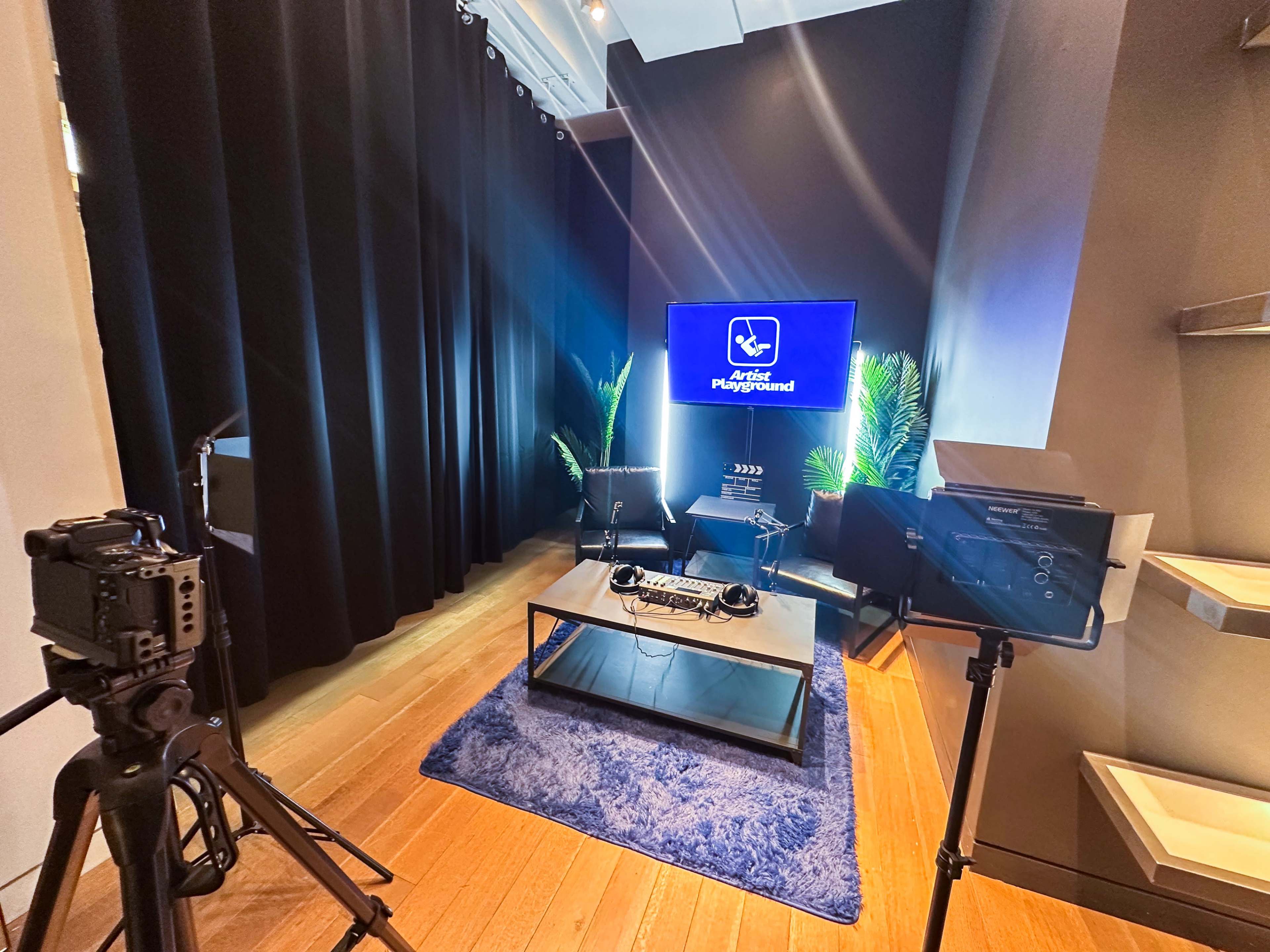 The image shows a small studio setup with a dark backdrop, a television displaying a logo, and equipment including cameras and microphones arranged on a table.