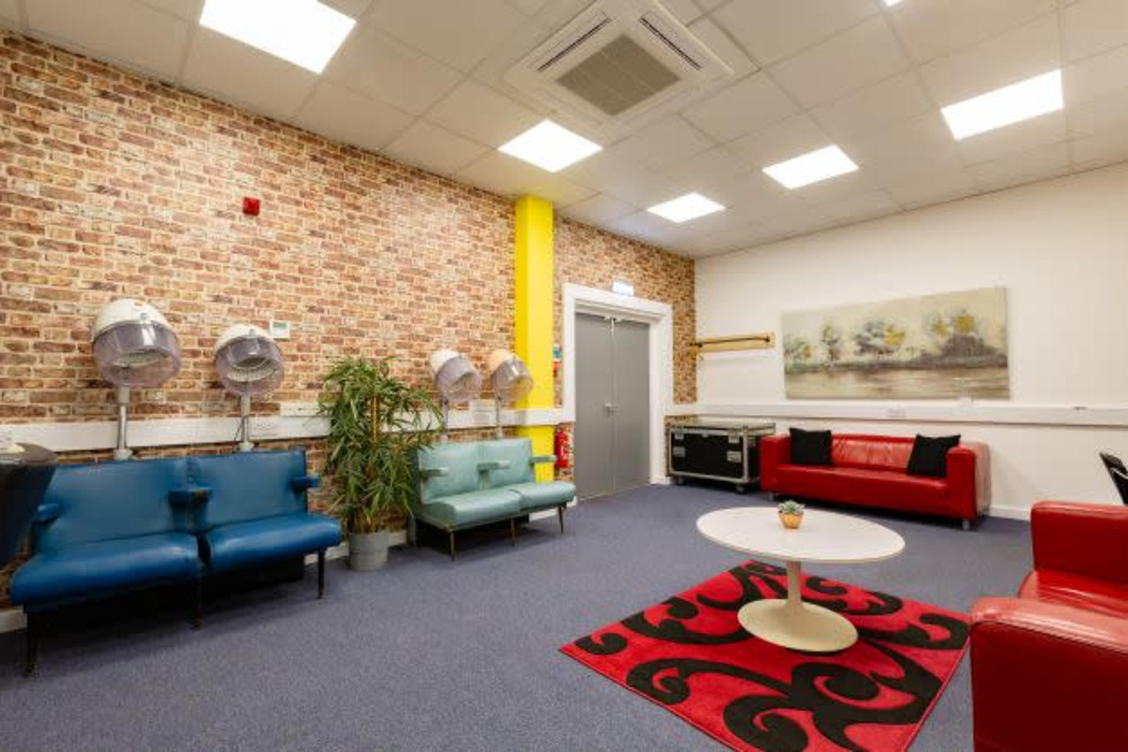A waiting room with brick walls, blue and red couches, a round white table, and hair drying stations on one side.