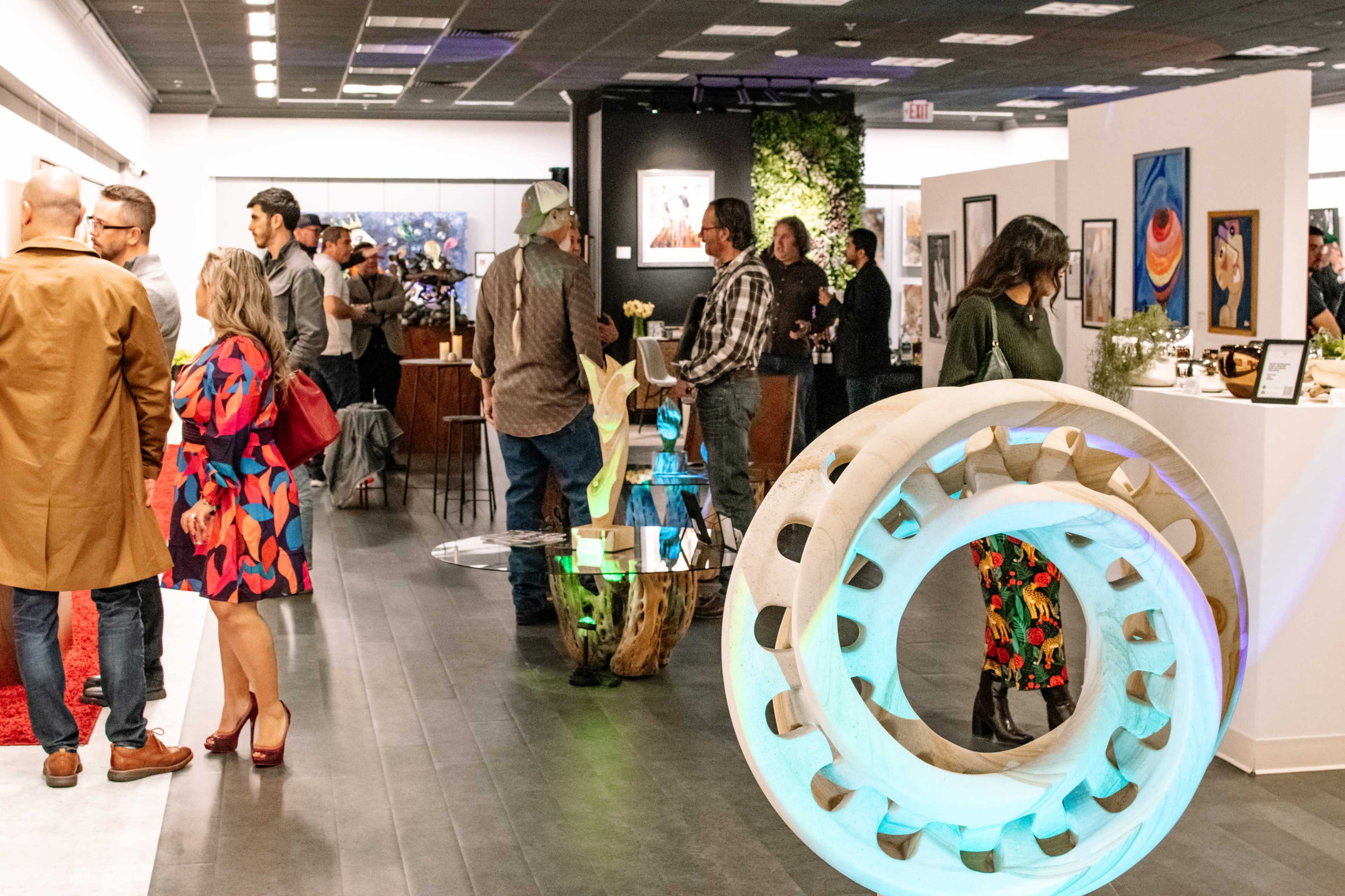 The image depicts a bustling art gallery event, featuring groups of people engaging in conversation among various artworks and installations.