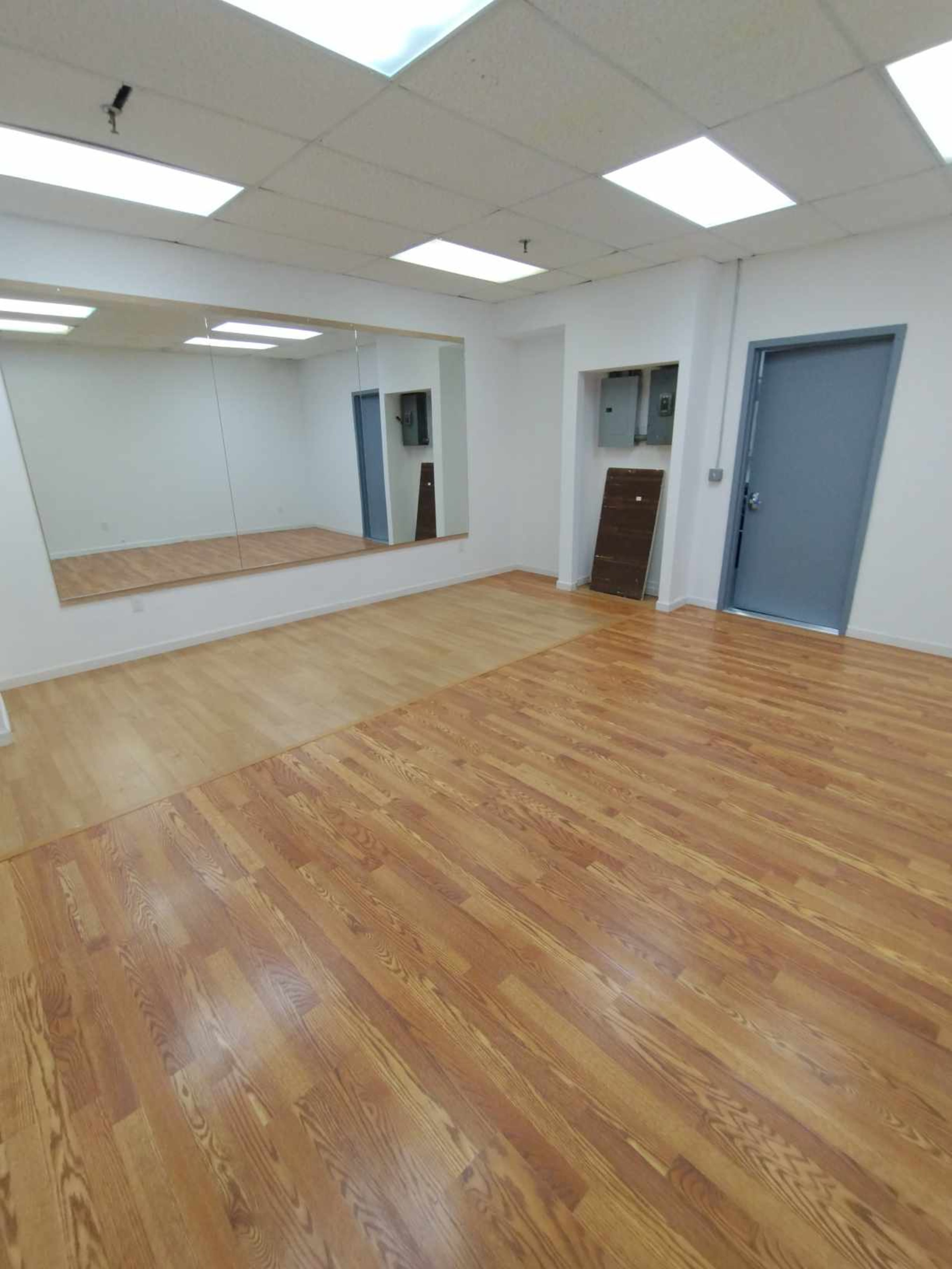 The image shows a spacious room with wooden flooring, white walls, a large mirror, and a door on one side.
