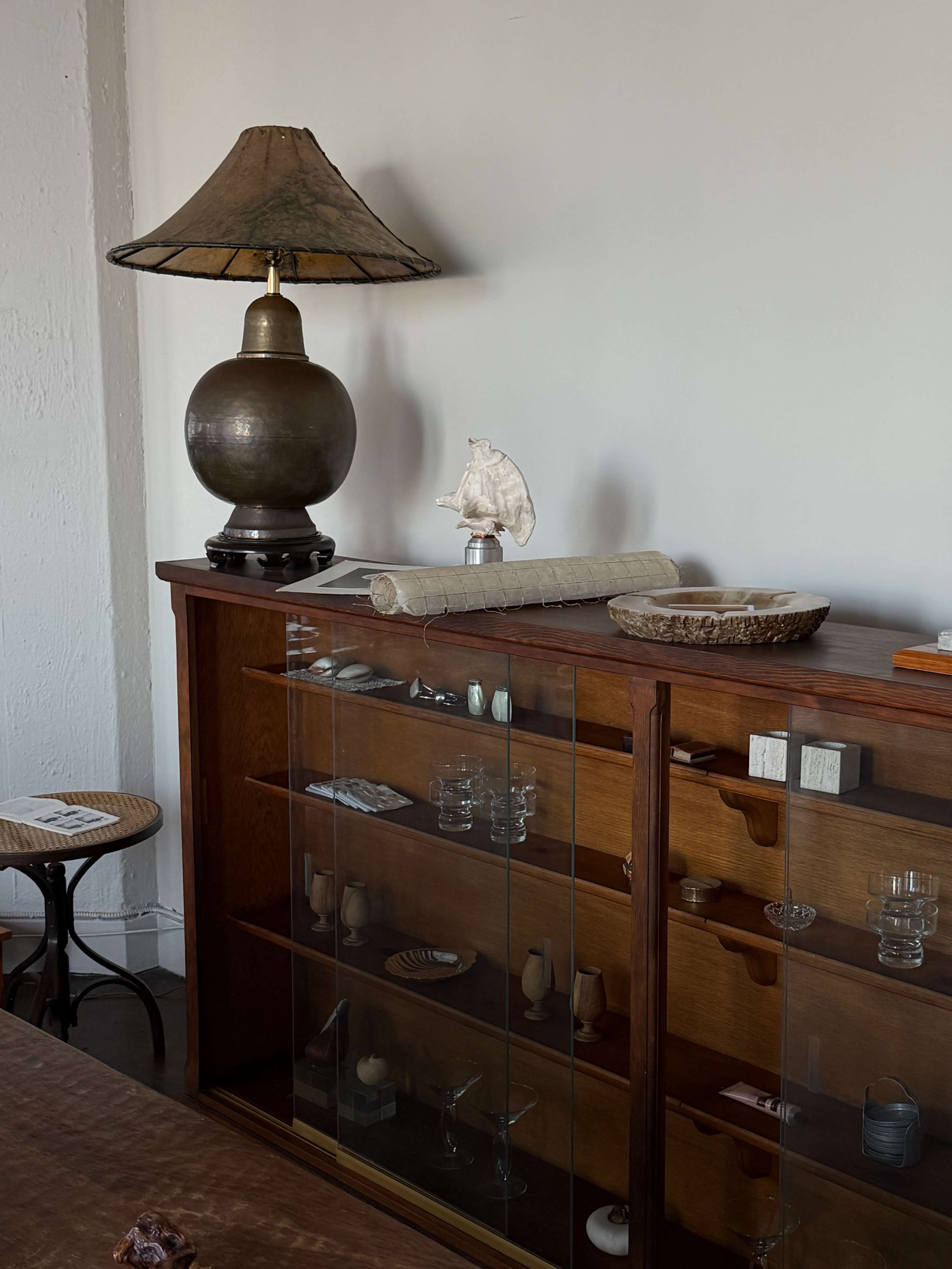 A wooden display cabinet with glass doors contains various small objects and decorations, next to a lamp with a round base and an alabaster figure of a bird.