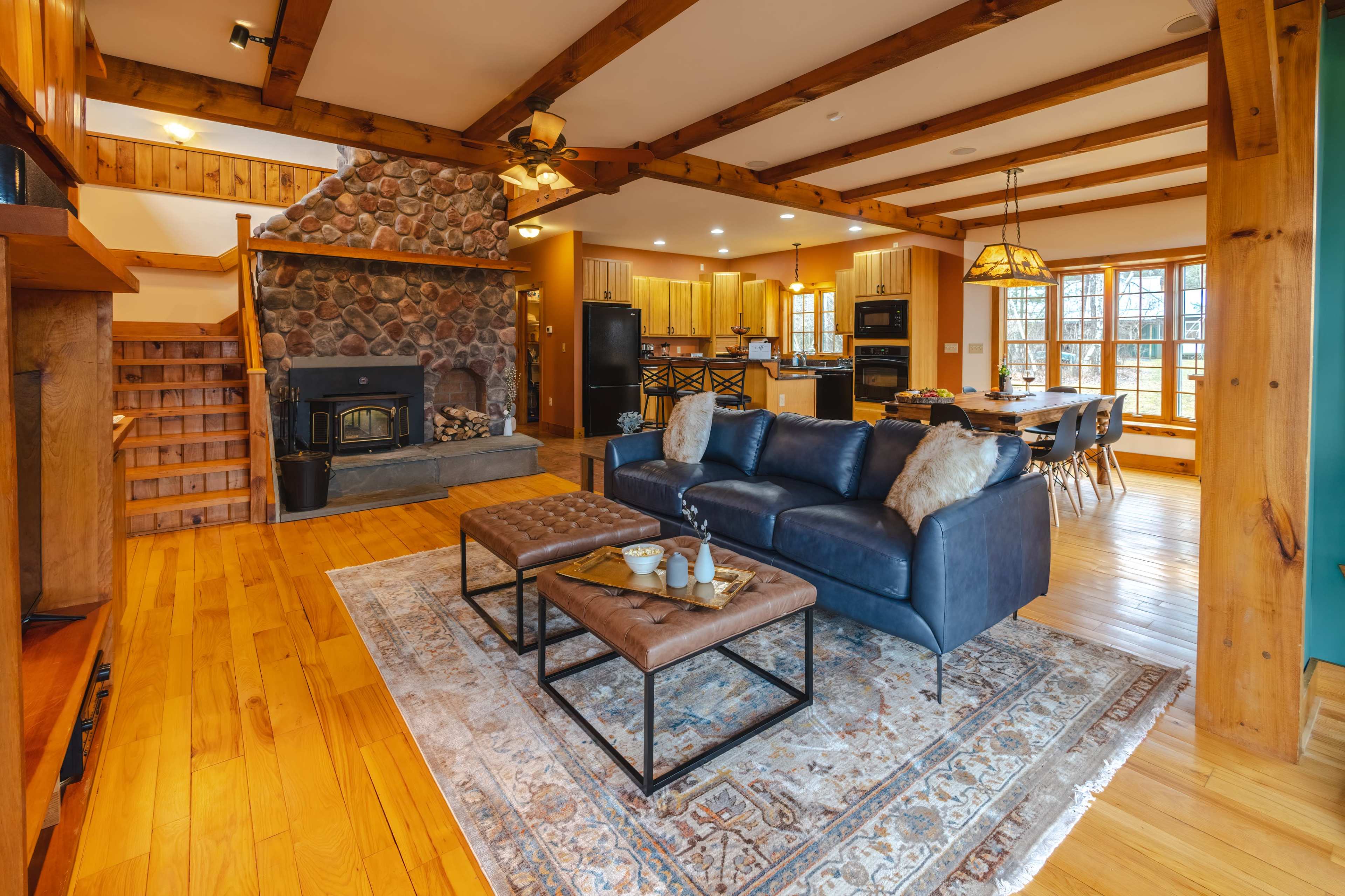A spacious living area with a stone fireplace, wooden beams overhead, a leather sofa, and a dining area in the background.