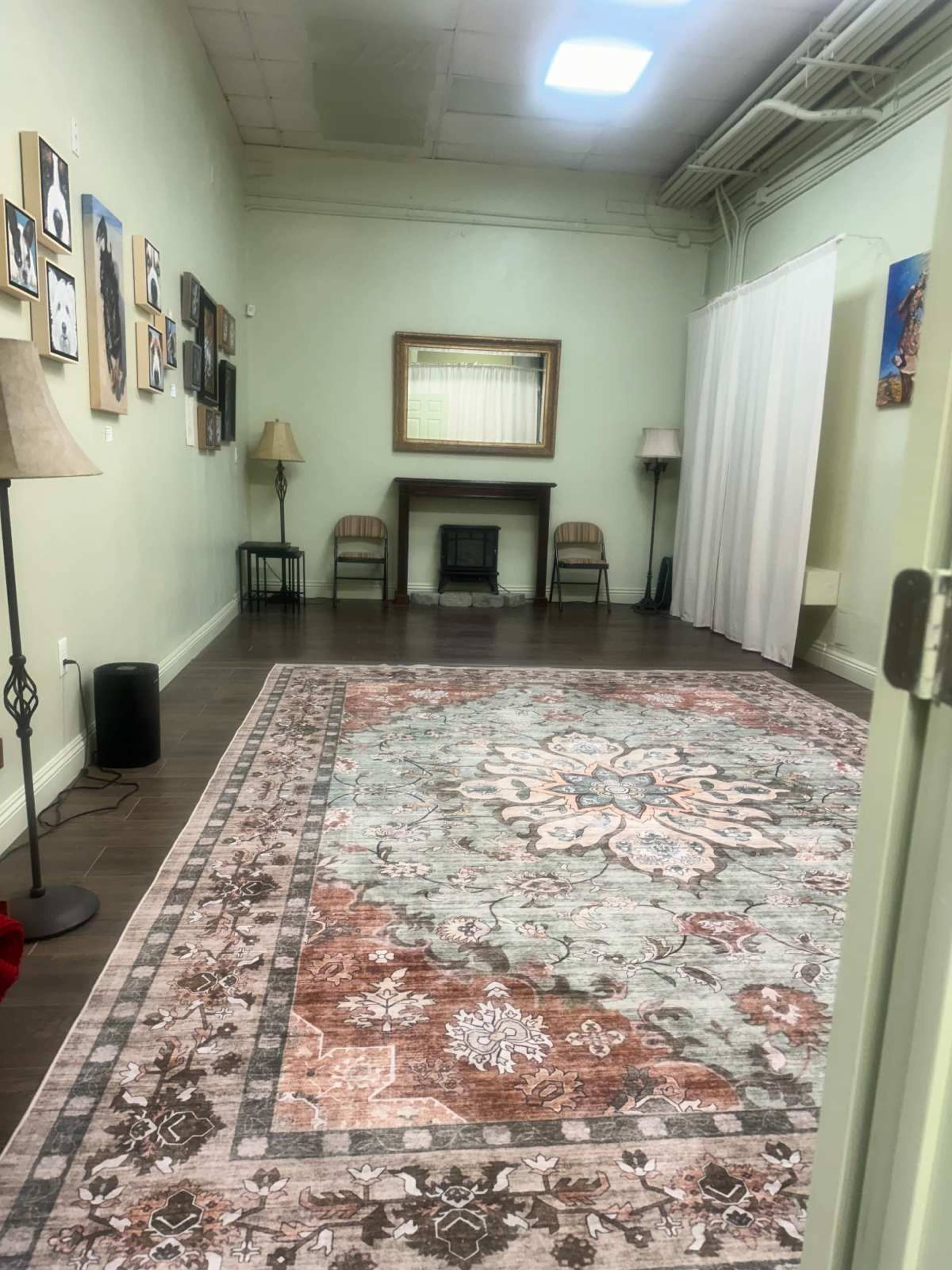 The image shows a spacious room featuring a large decorative rug, two chairs, a fireplace, wall art, and light-colored walls.