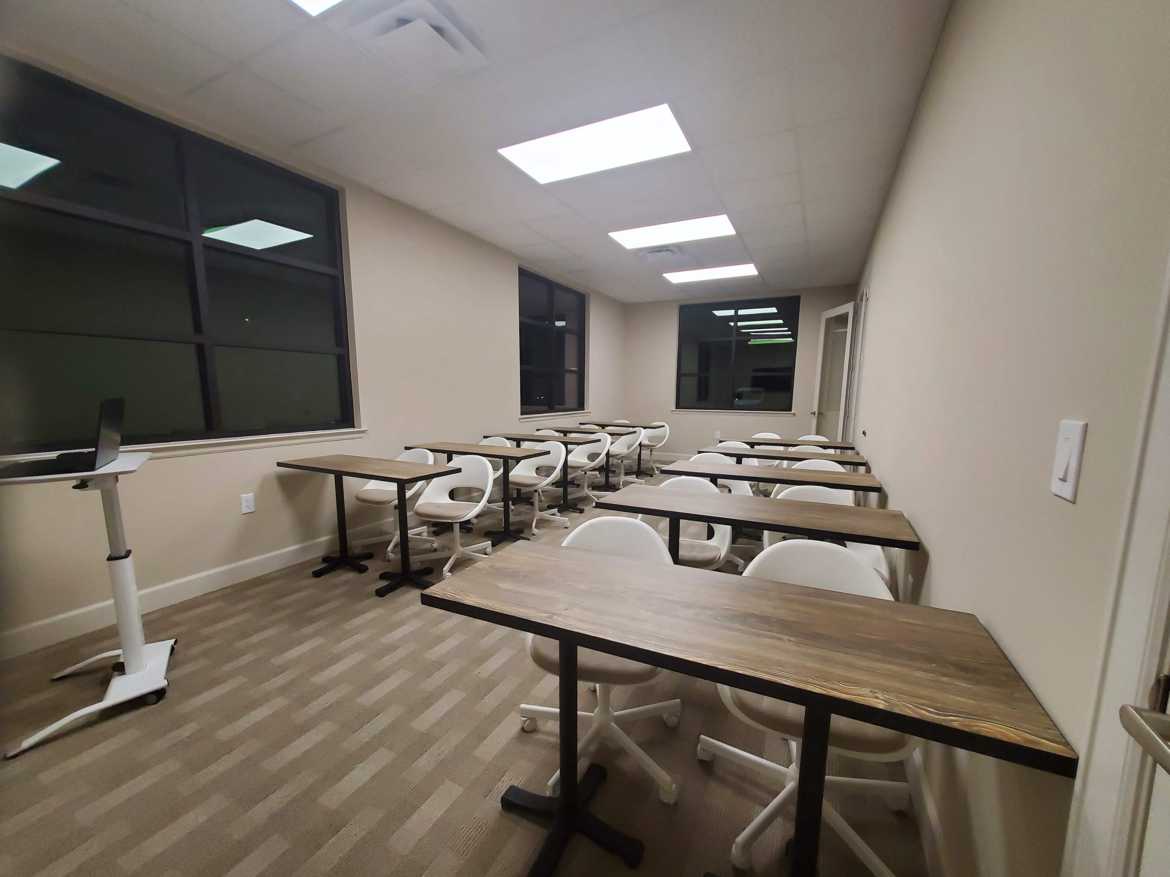 The image shows a classroom with several empty tables and chairs arranged in rows, illuminated by overhead lights and natural light from large windows.