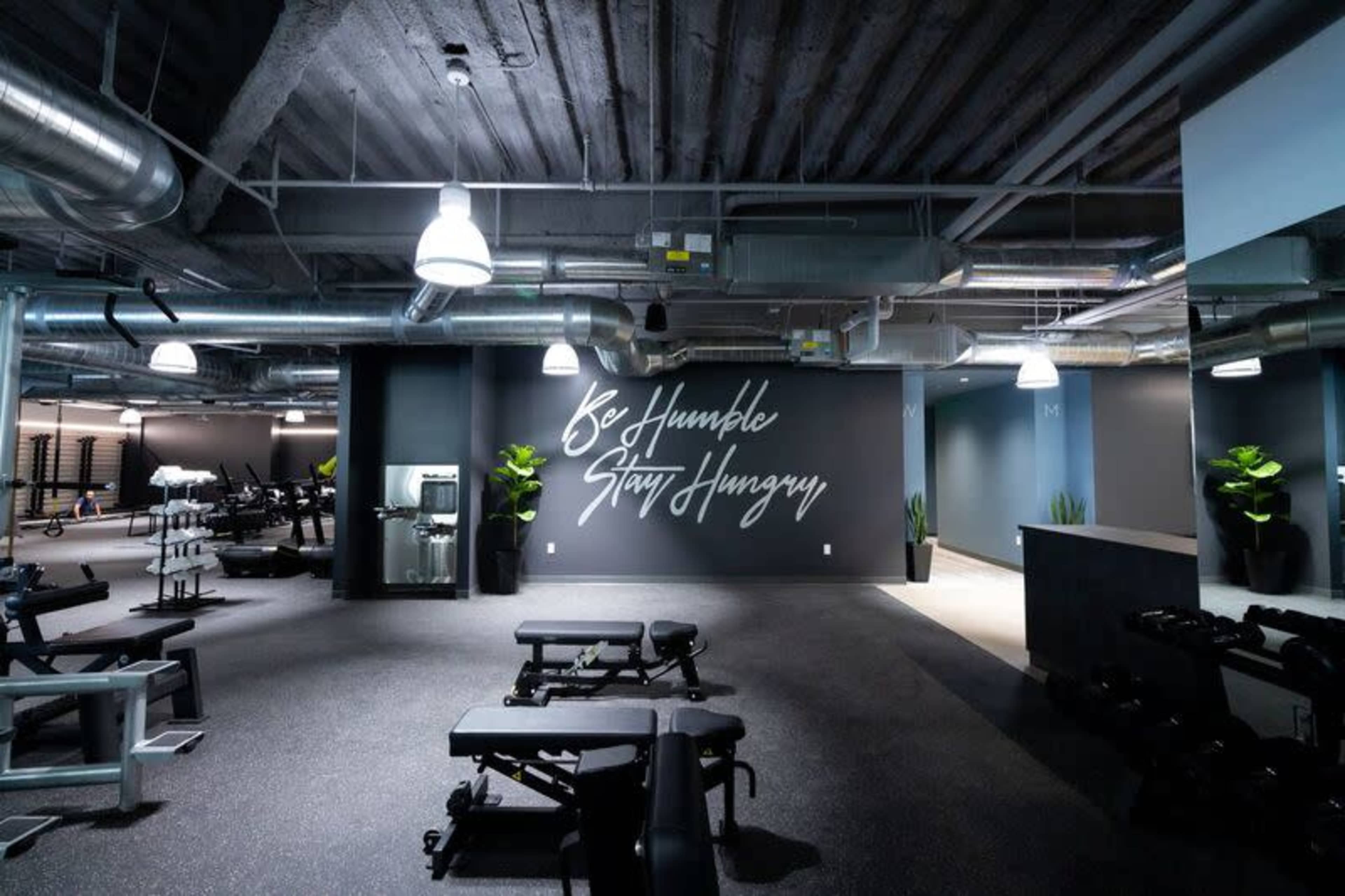 The image shows a modern gym interior with workout benches, free weights, and a motivational wall mural that reads "Be Humble, Stay Hungry."