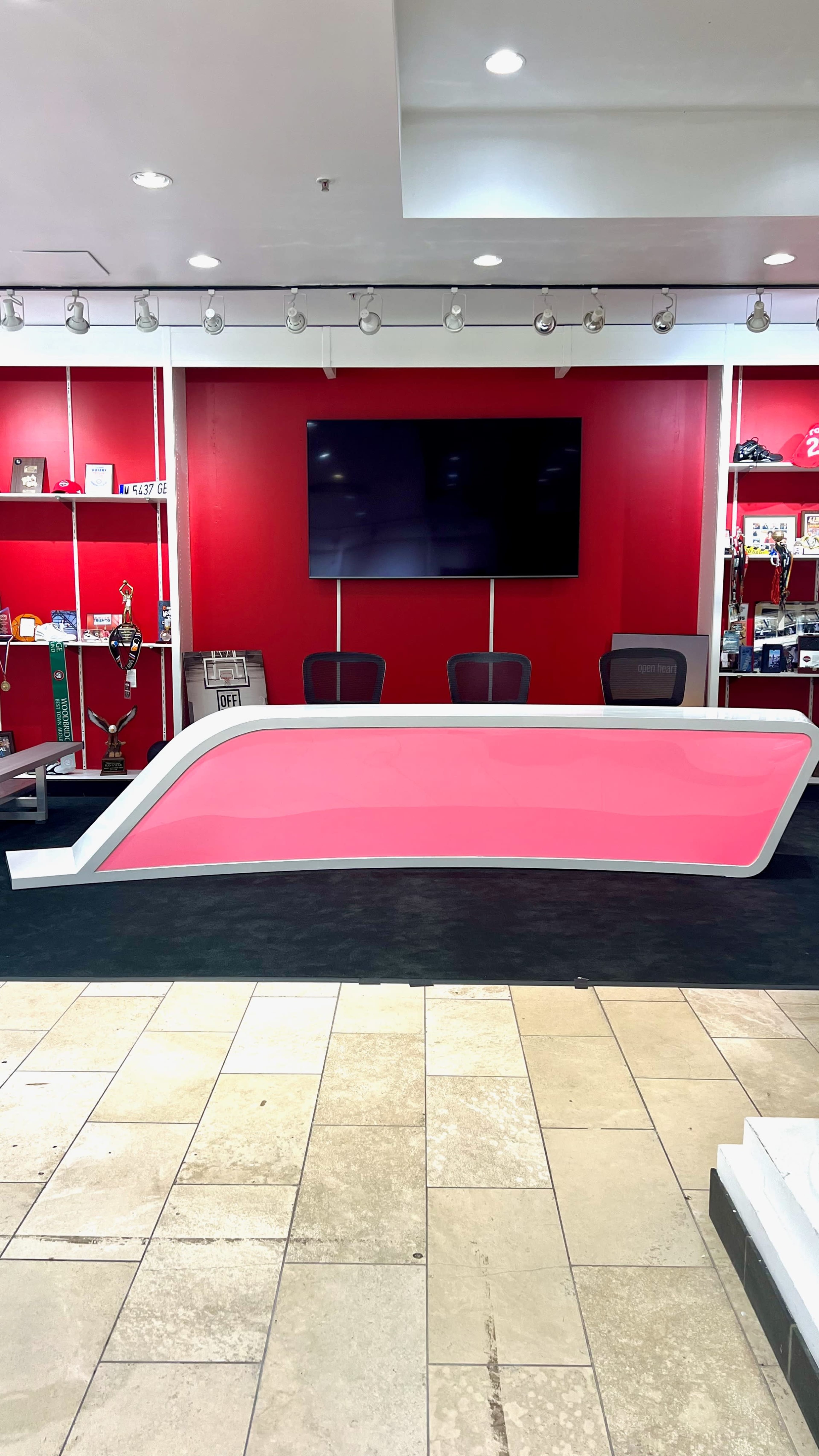 The image shows a modern reception desk with a pink front, positioned in front of a large television screen and against a red wall, in a well-lit space with tile flooring.