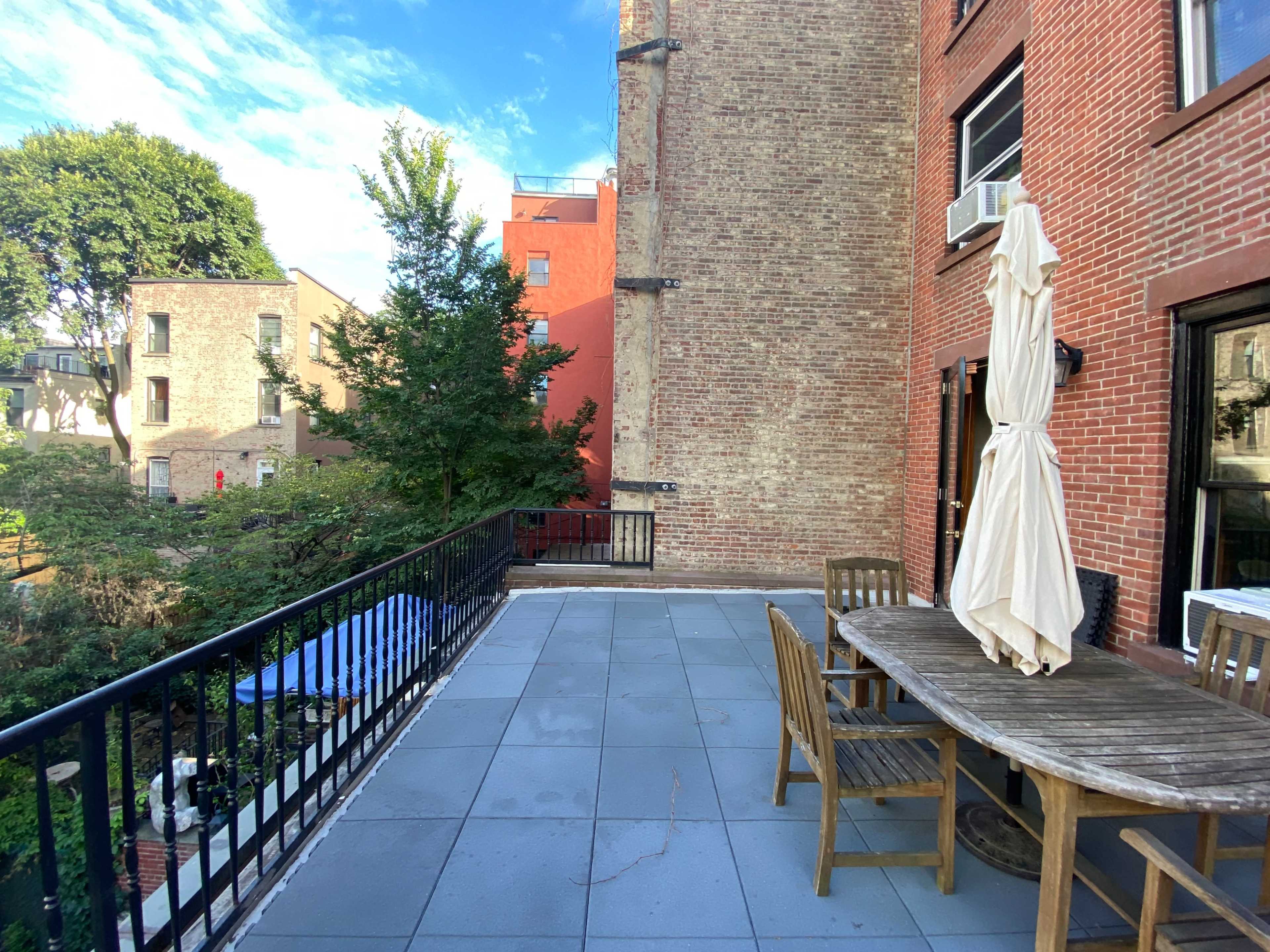 A spacious patio area with a wooden table and chairs, surrounded by brick walls and trees, overlooking a green space.