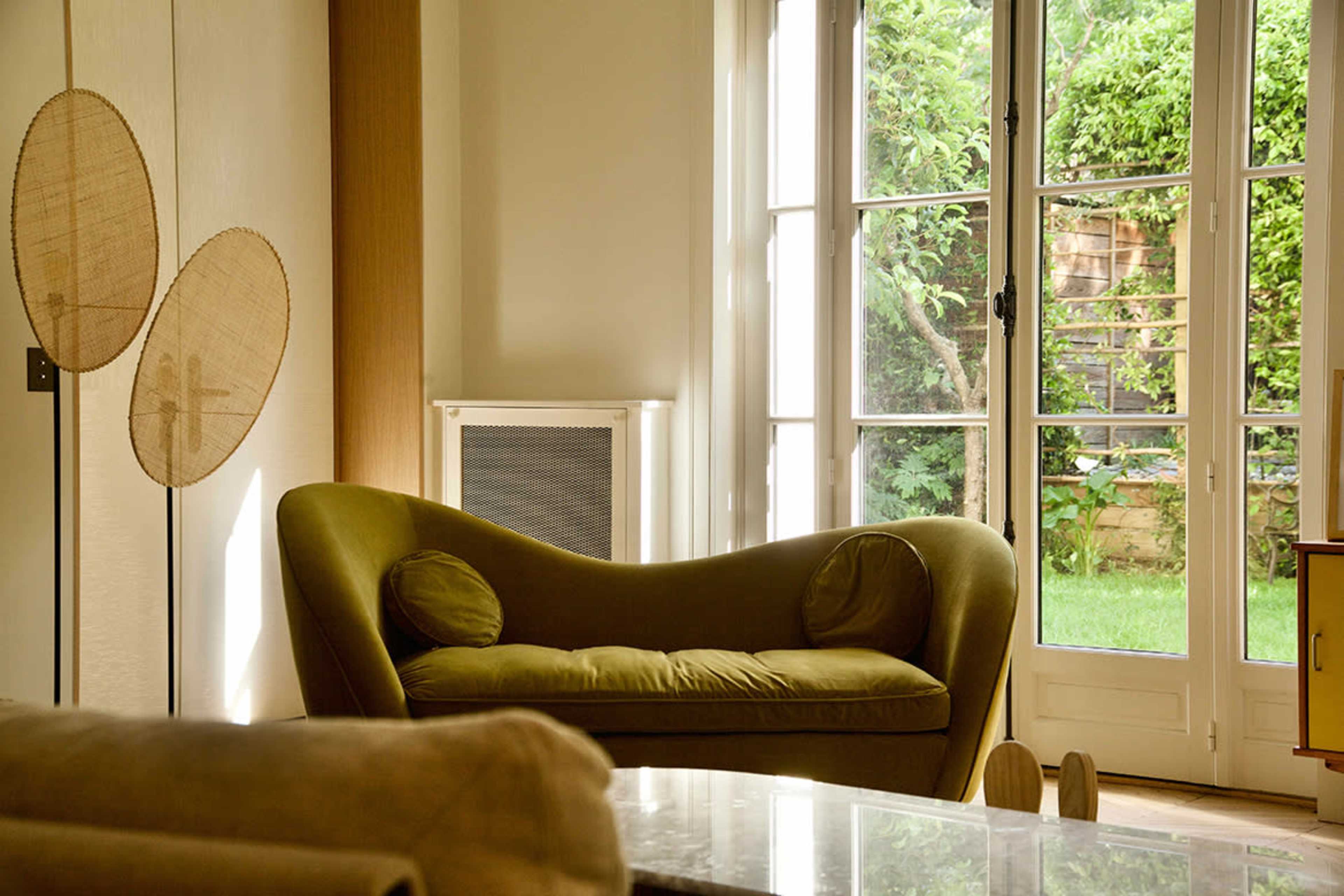 The image shows a cozy living room featuring a green curved sofa positioned near large windows overlooking a garden.