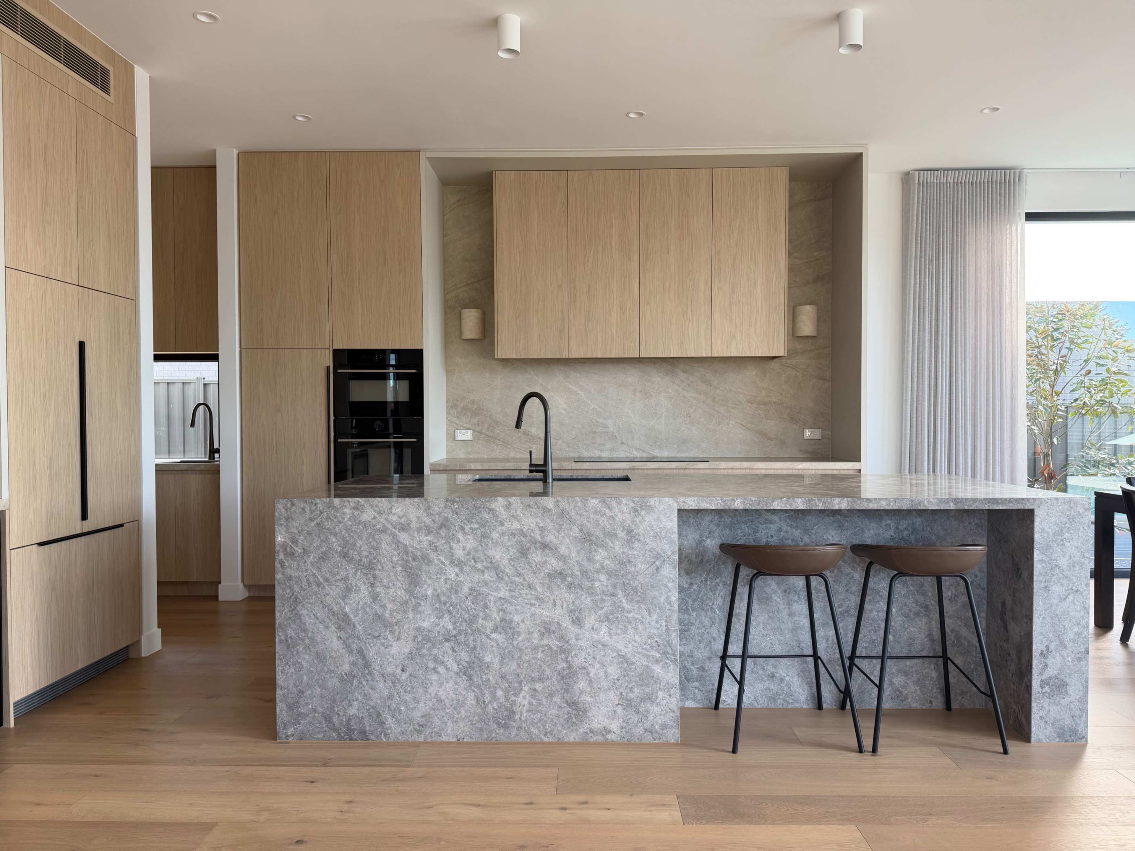 The image shows a modern kitchen featuring a gray stone countertop, wooden cabinetry, and two stools at a kitchen island.