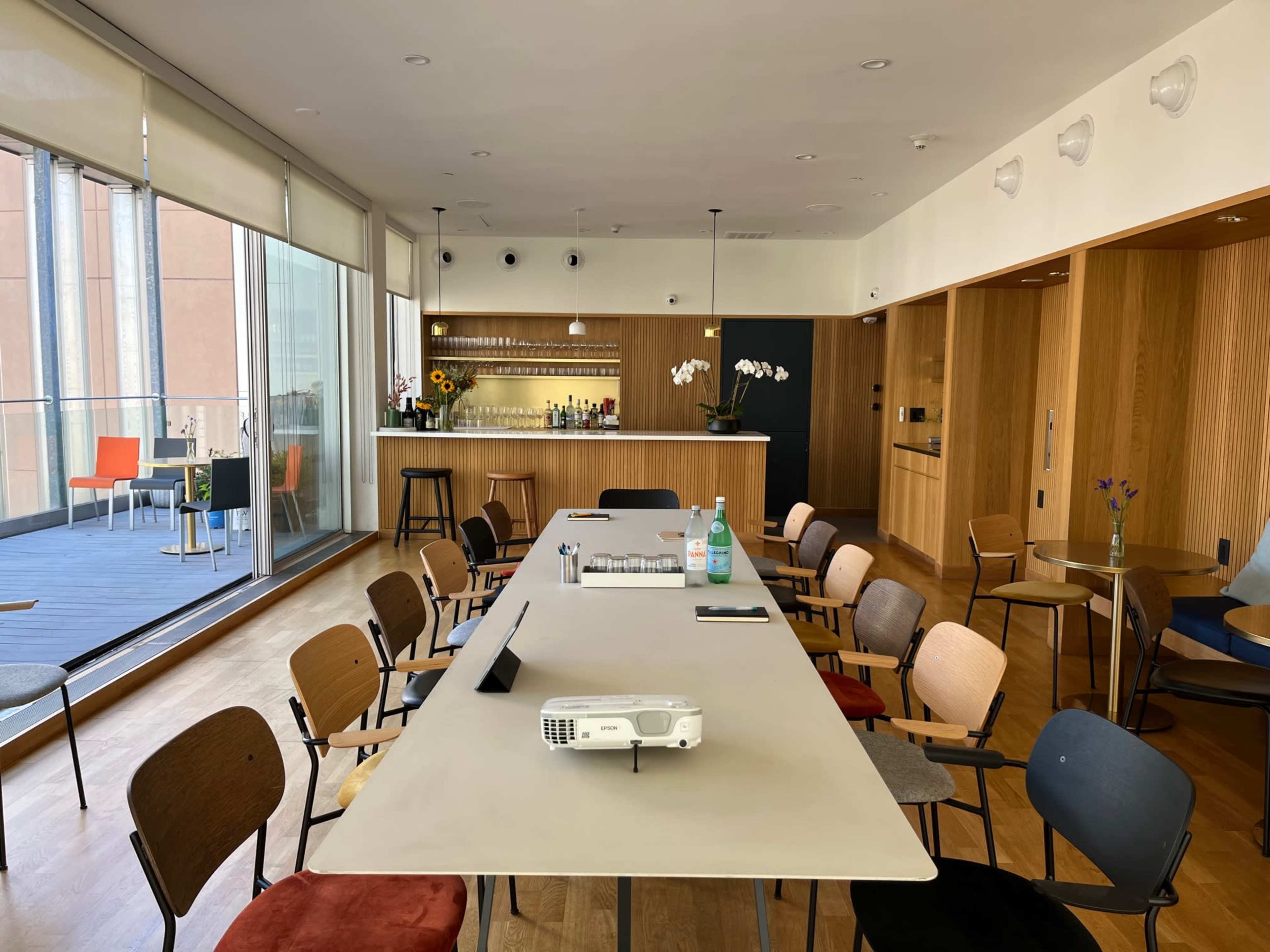 A bright interior features a long table surrounded by various chairs, with a bar area in the background and large windows letting in natural light.