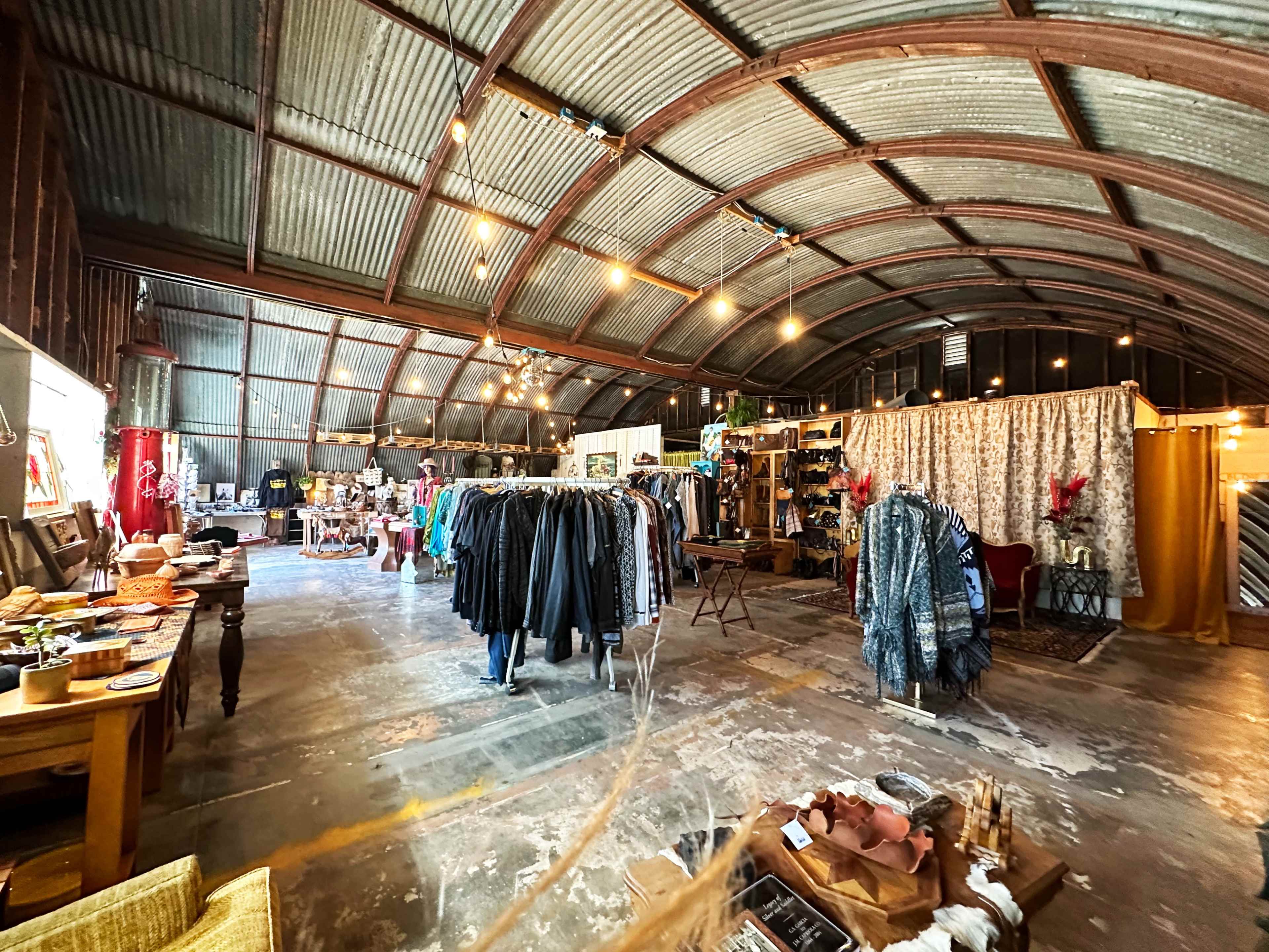 A spacious, industrial-style interior of a vintage store featuring clothing racks, wooden tables with decorative items, and various displays under soft lighting.