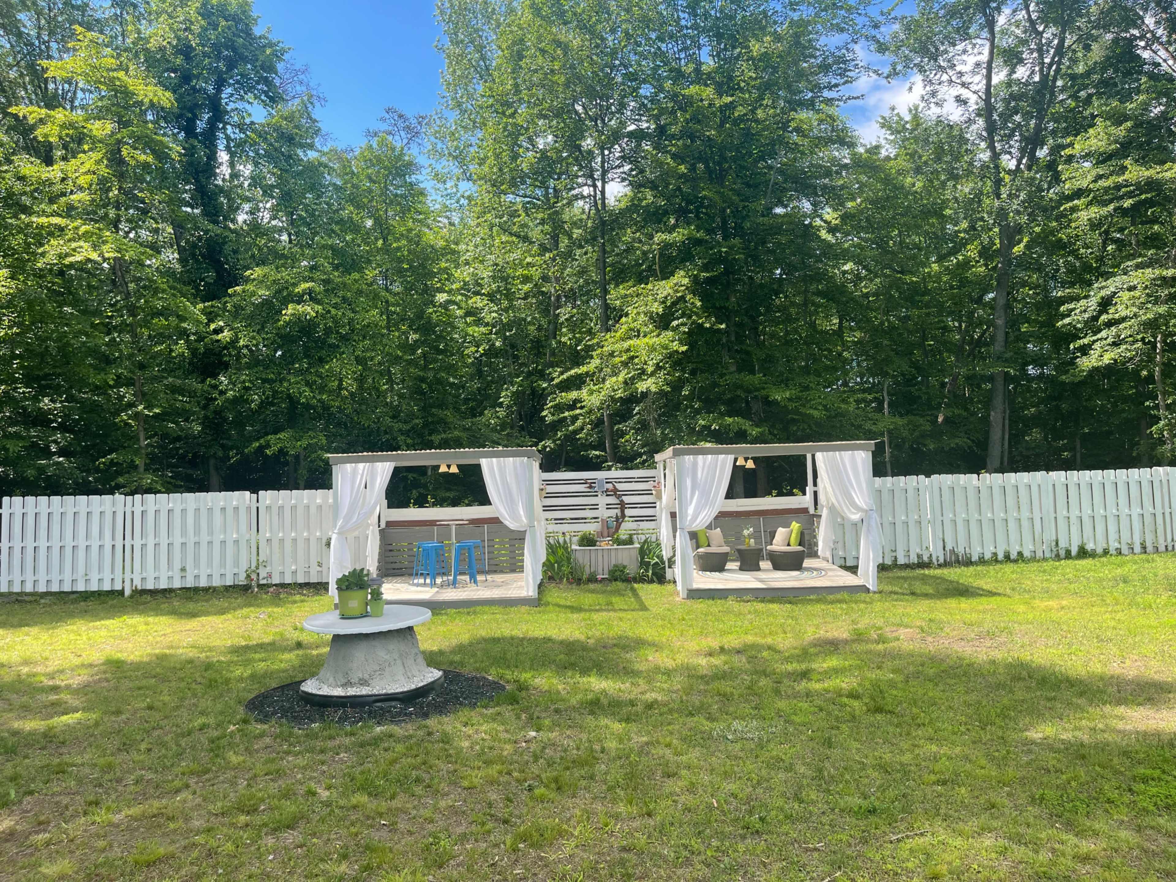 The image shows a backyard with two open pavilions separated by a circular stone table, surrounded by a white fence and lush greenery.
