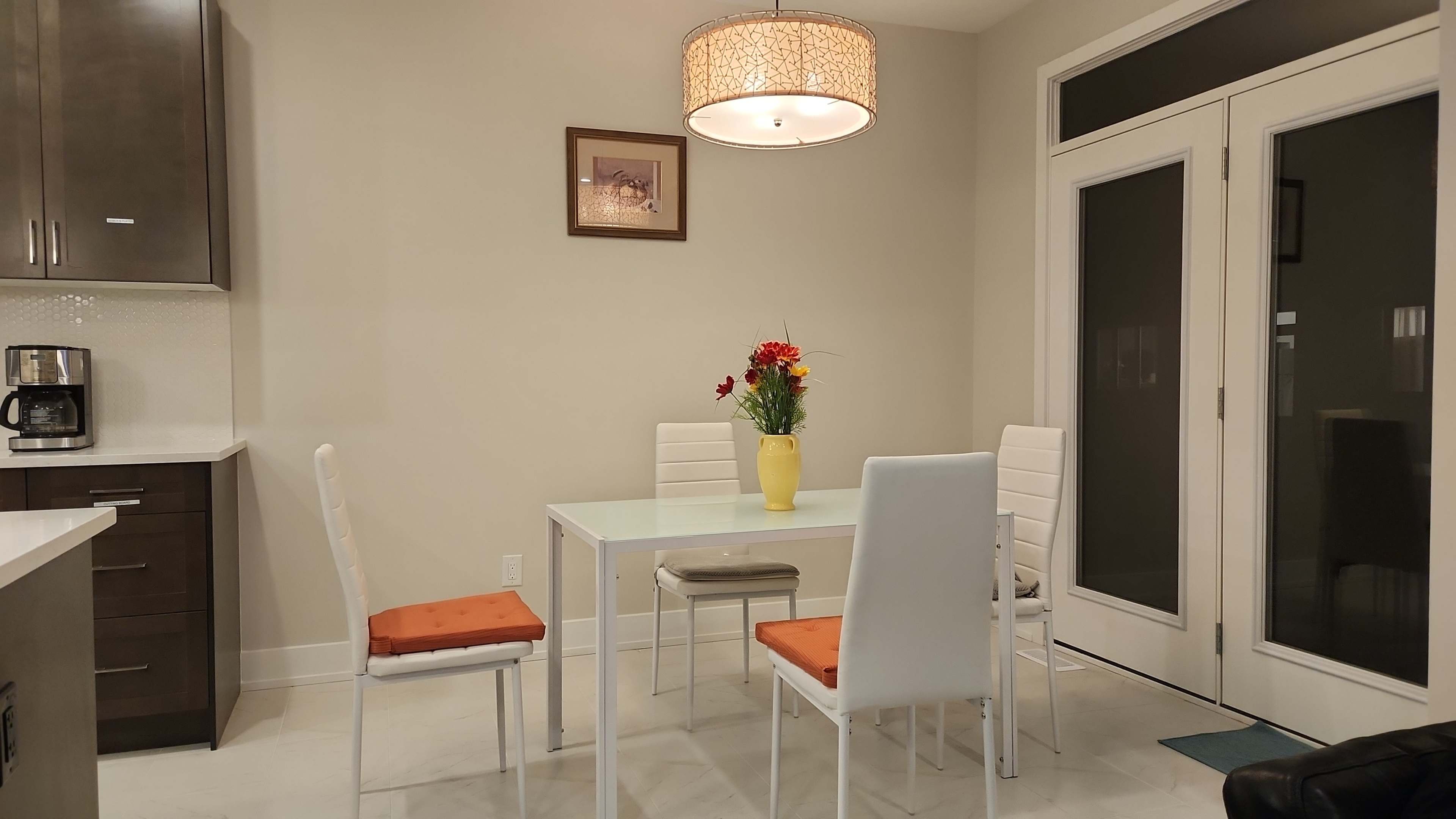 A modern dining area features a white table surrounded by four chairs, with a vase of flowers at the center and a large light fixture overhead.