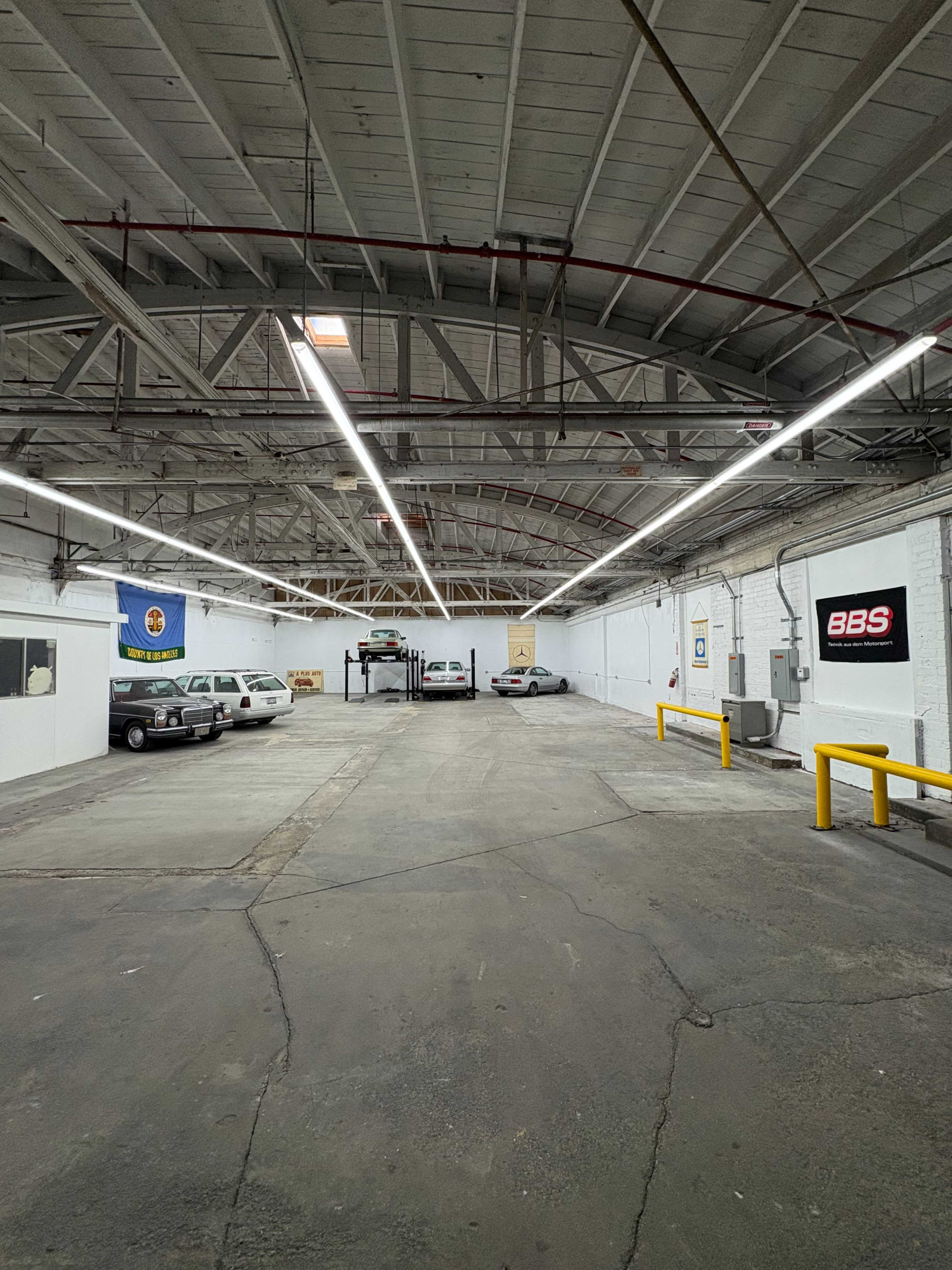 The image shows an empty garage or workshop space with several vehicles parked, high ceilings, and overhead lighting.