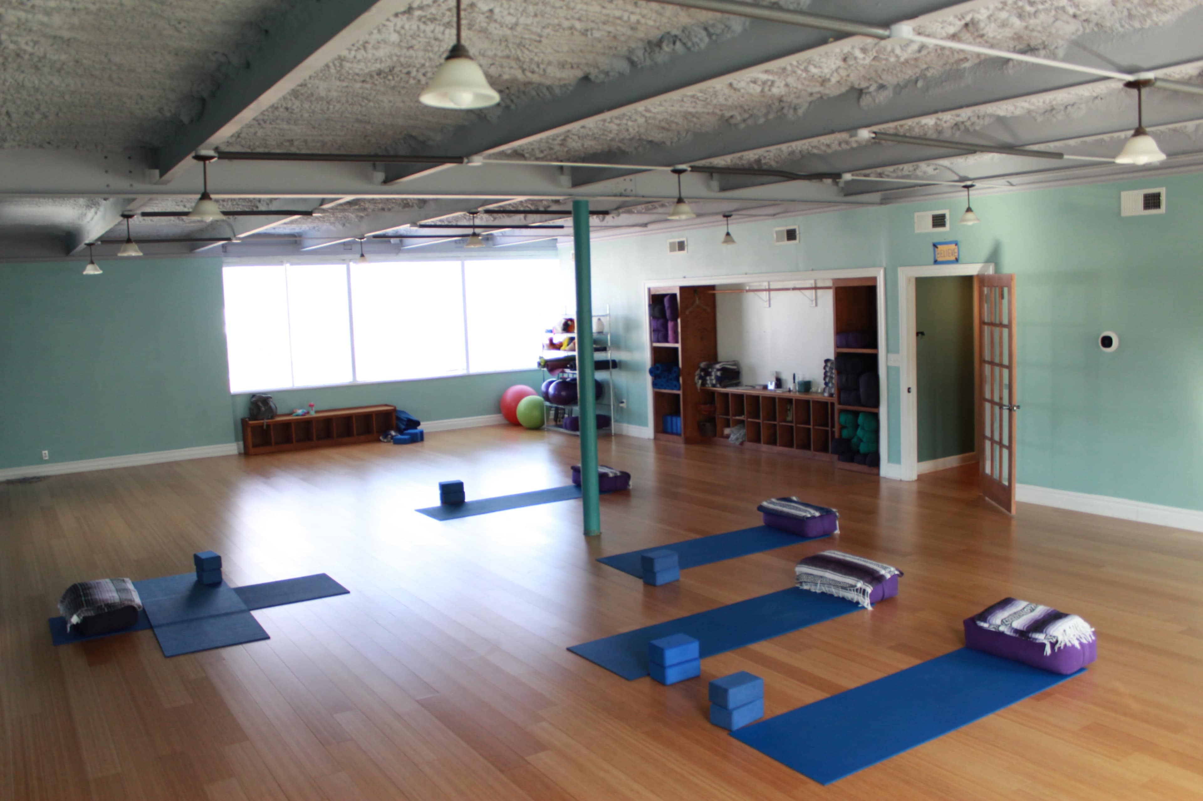 The image shows a spacious yoga studio with wooden floors, blue mats arranged on the floor, and various props such as blocks and bolsters neatly placed.