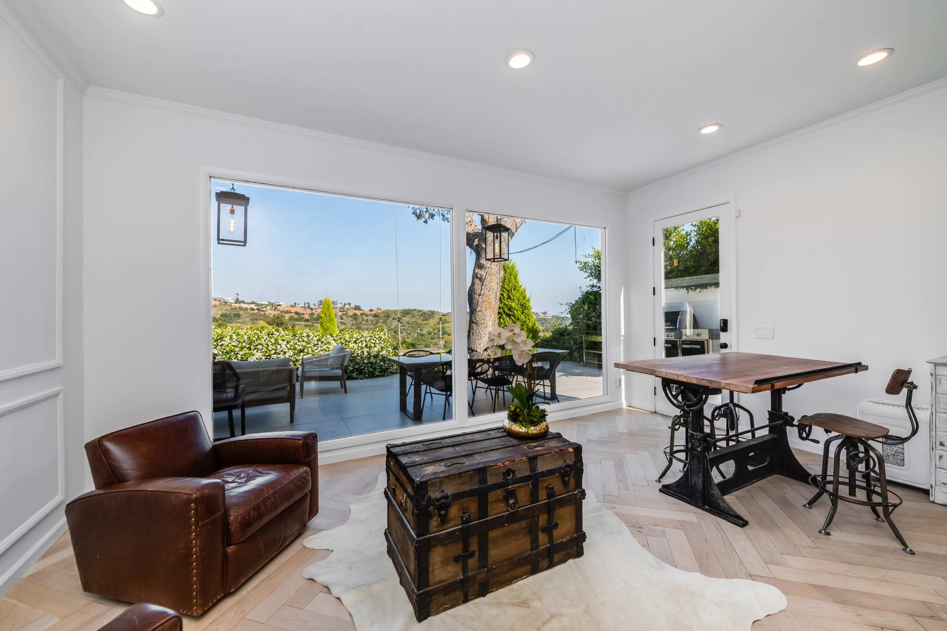 A well-lit room features a leather armchair, a wooden table, and a vintage trunk, with large windows offering a view of an outdoor garden.