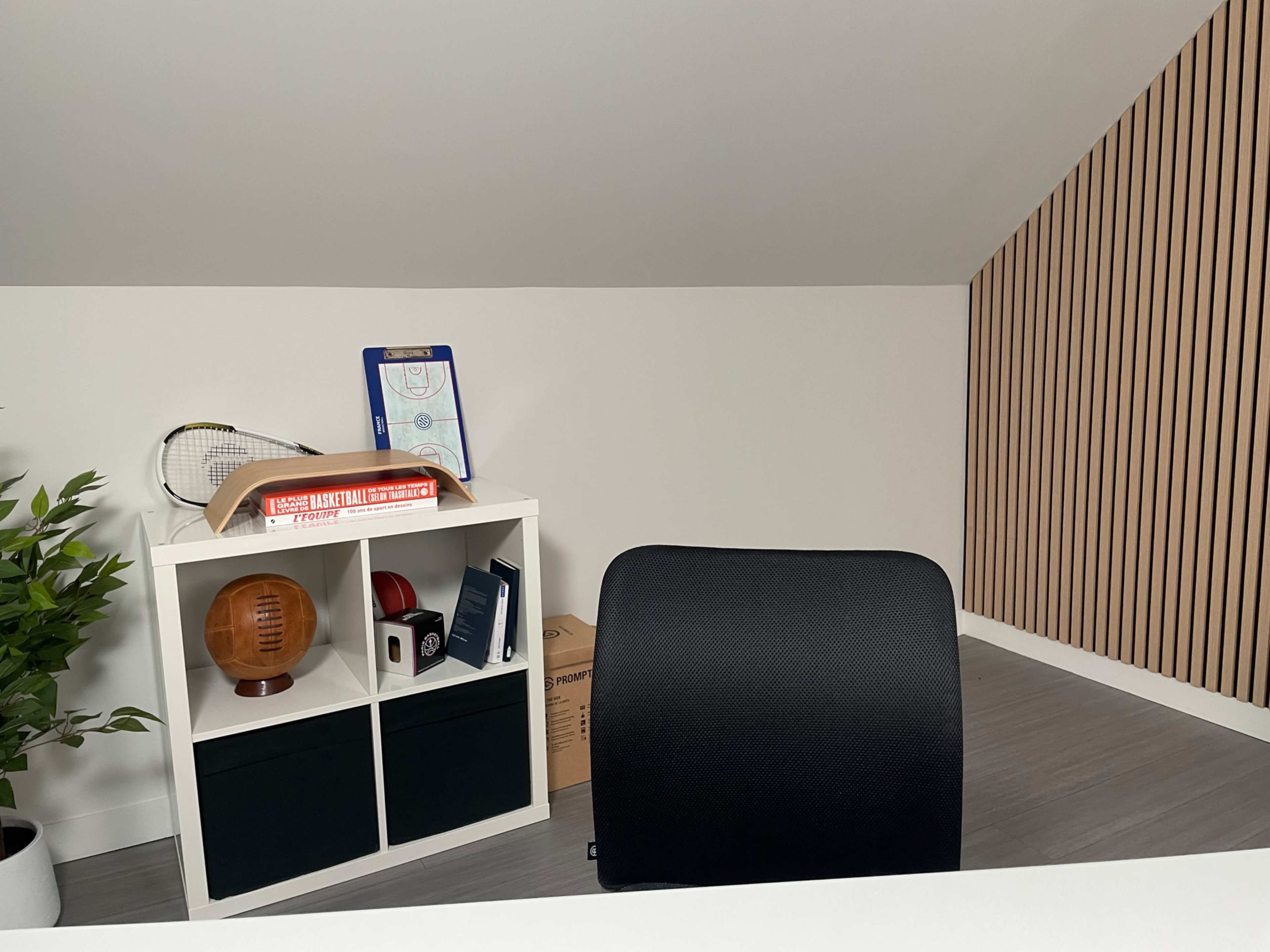 The image shows a minimalist home office with a small bookshelf containing sports memorabilia and a clipboard against a backdrop of slanted walls and wooden panels.