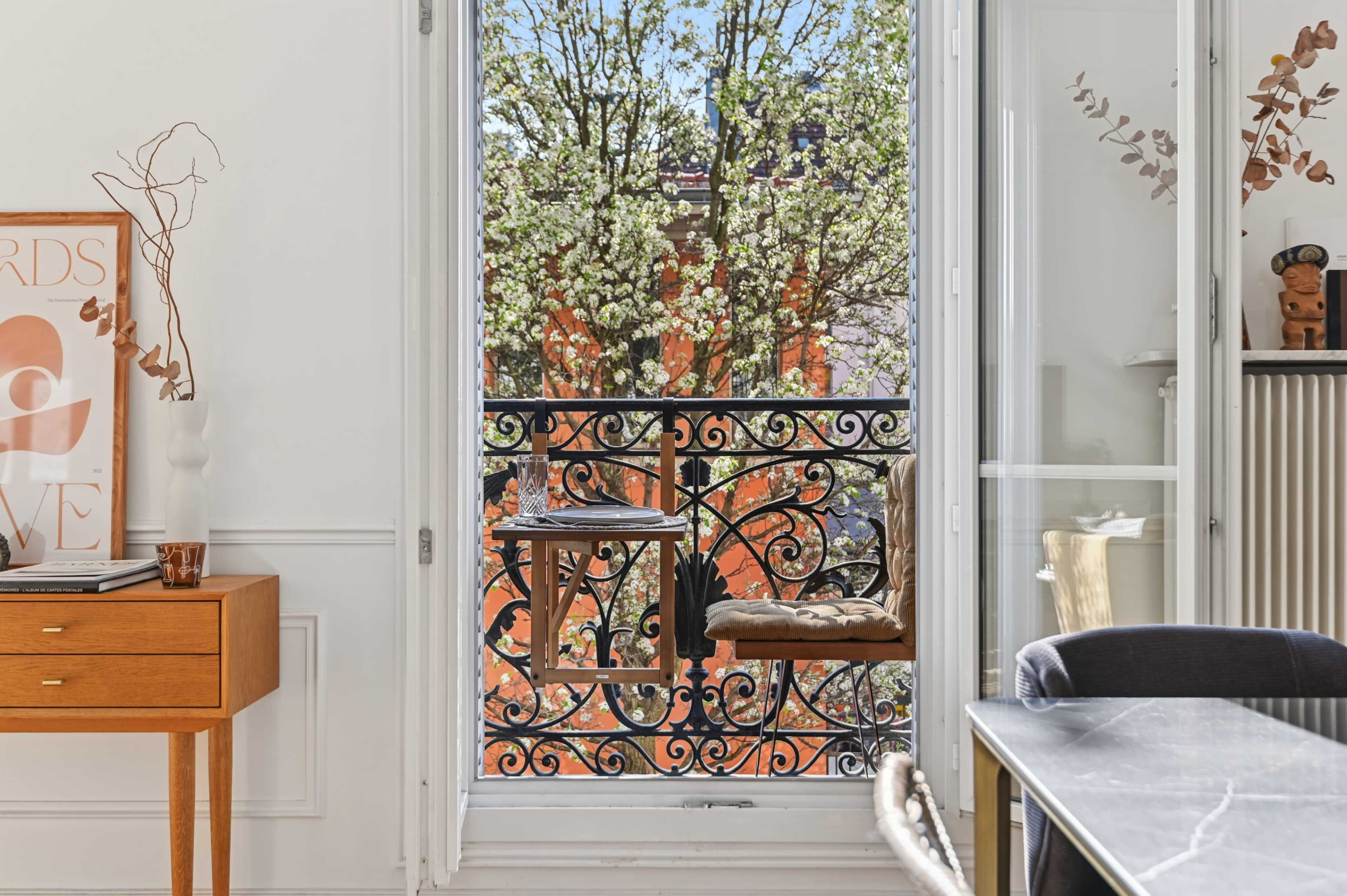 A balcony with an ornate iron railing overlooks a tree in bloom, viewed from a well-furnished interior space.