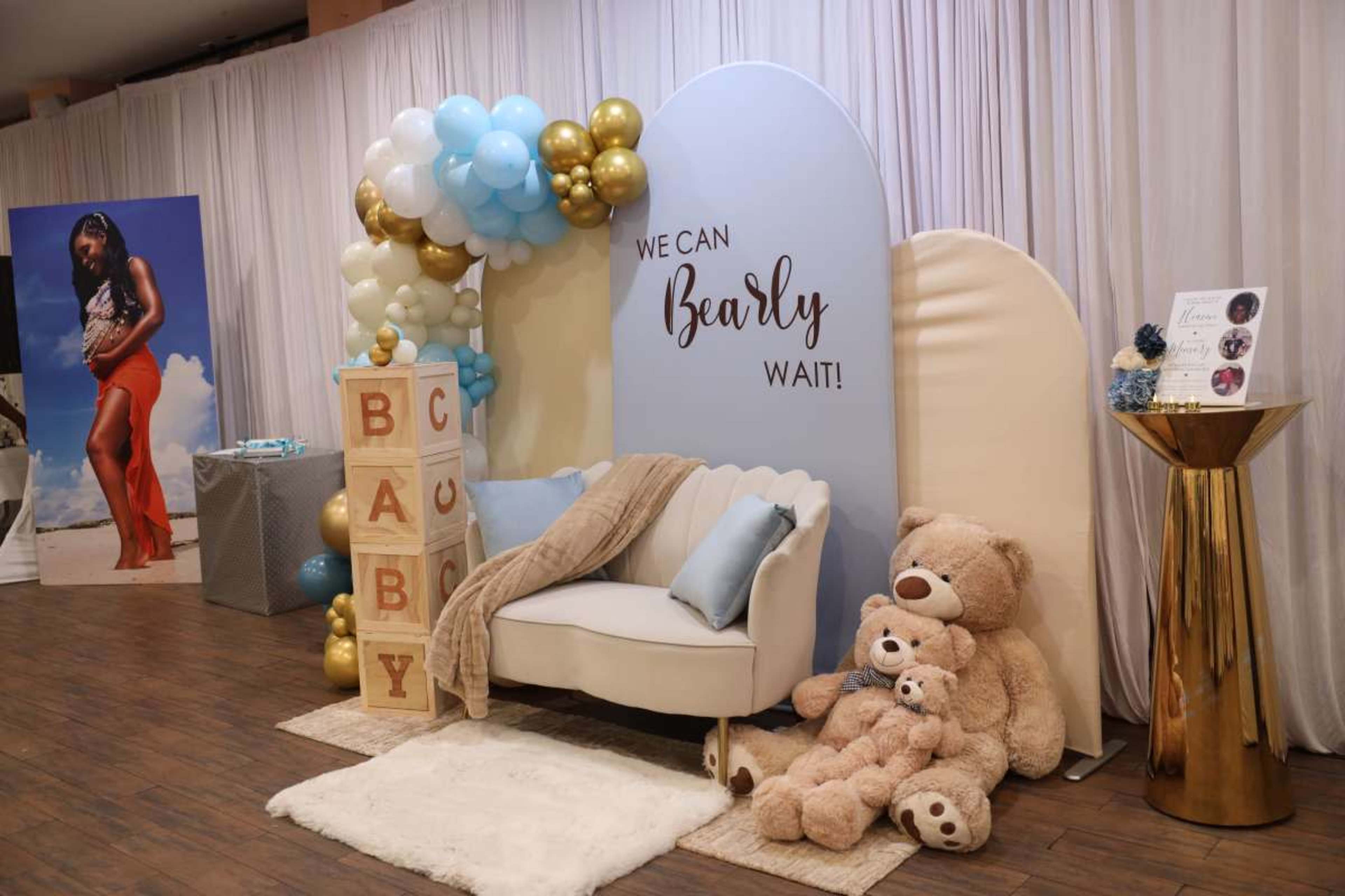 The image features a decorative baby shower setup with a couch, teddy bears, balloon arch, and a sign reading "WE CAN BEARLY WAIT!" against a backdrop of soft drapes.