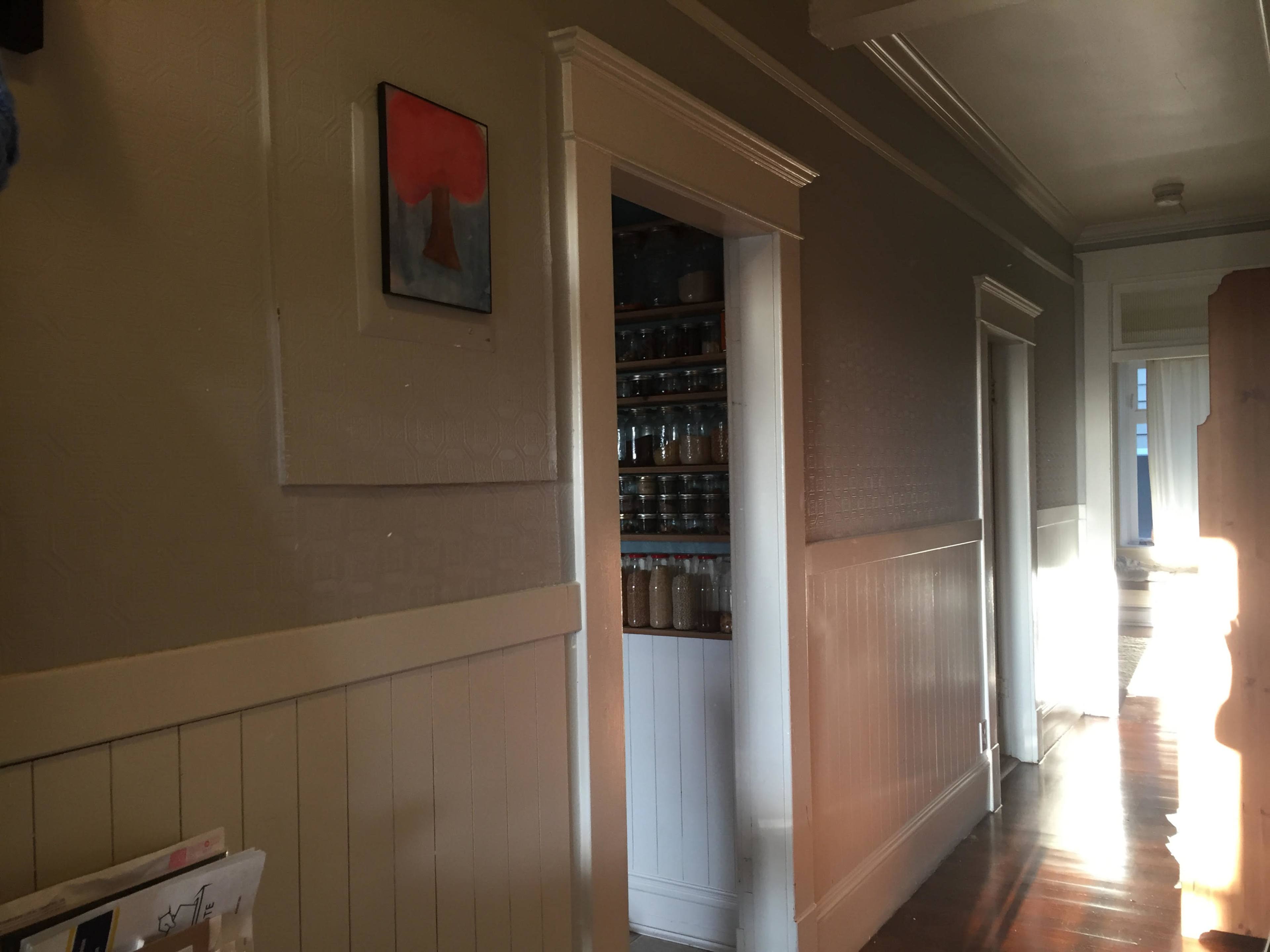 The image shows a hallway with light streaming in, featuring a wall with a painting and a doorway leading to a space with shelves filled with jars.