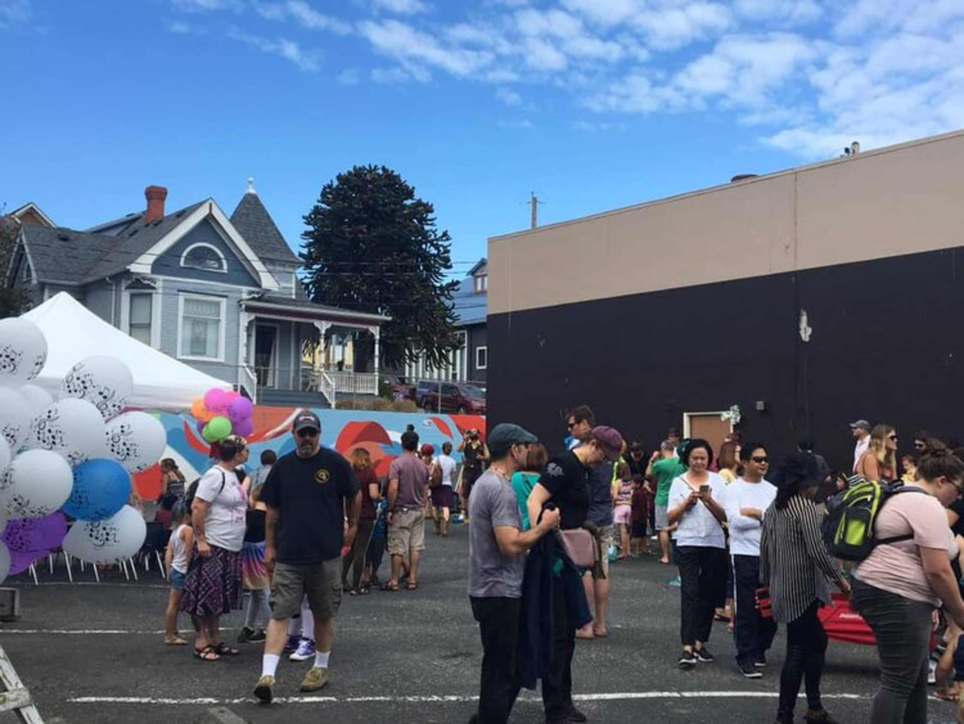 A large crowd gathers in a parking lot for an outdoor event, with a colorful mural on one wall and a white tent set up nearby.