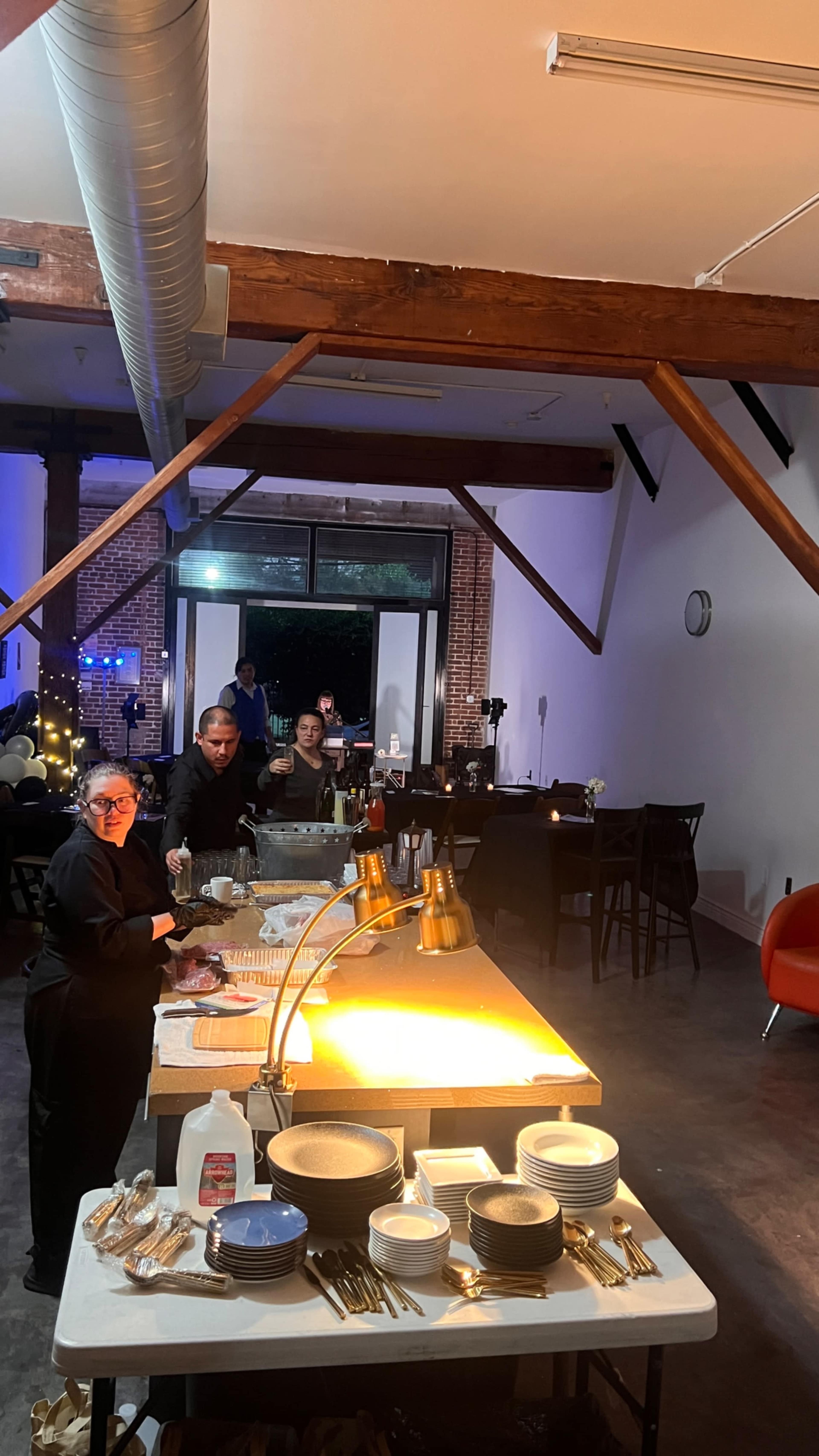 A buffet setup is shown in a well-lit event space, with two staff members preparing food as guests are seated in the background.