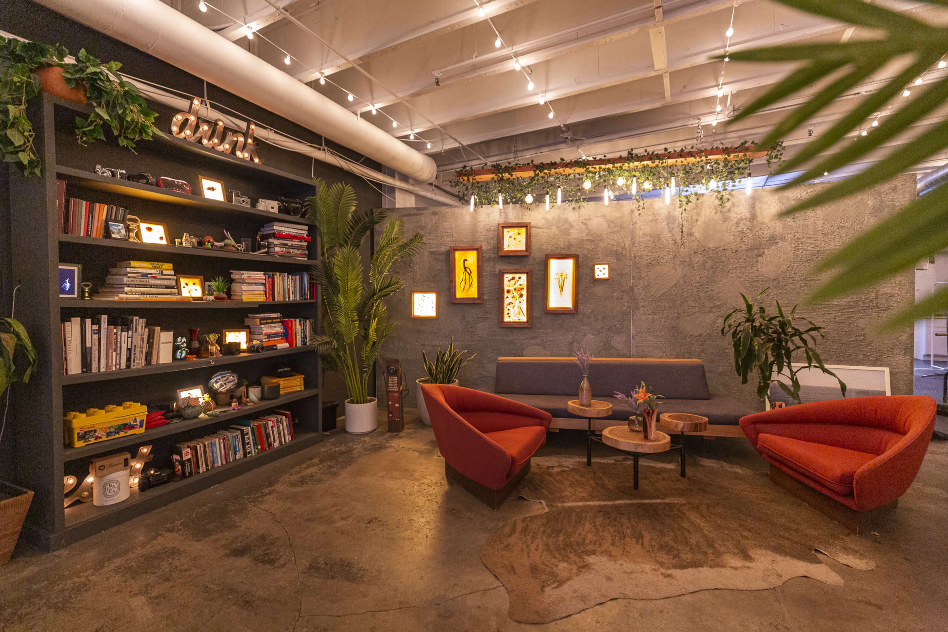 The image shows a cozy lounge area with two orange armchairs, a gray sofa, a bookshelf filled with books and decorative items, and plants, all under soft lighting from string lights.