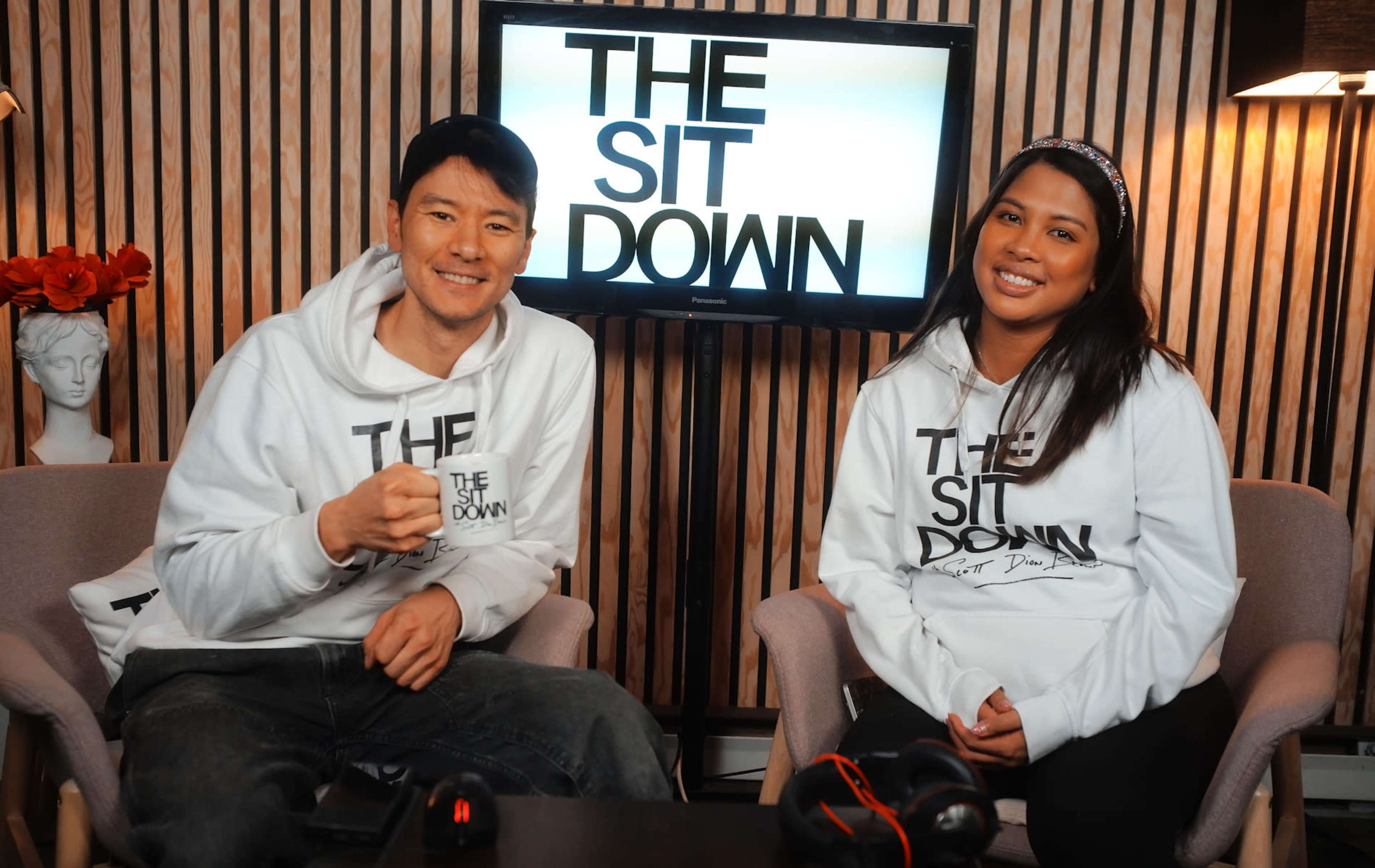 Two hosts wearing matching hoodies with "THE SIT DOWN" printed on them sit in a studio setting with a television displaying the same text behind them.