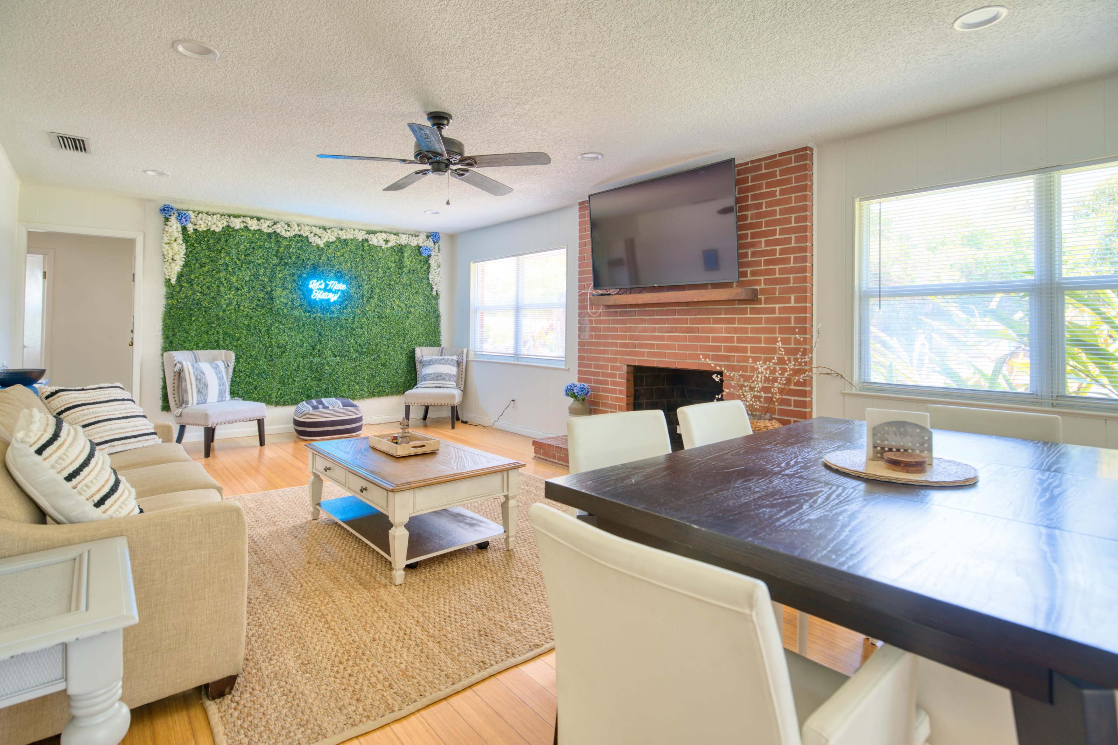 A living room with a green wall featuring decorative plants, a brick fireplace, a television, a sofa, and a dining table with chairs.