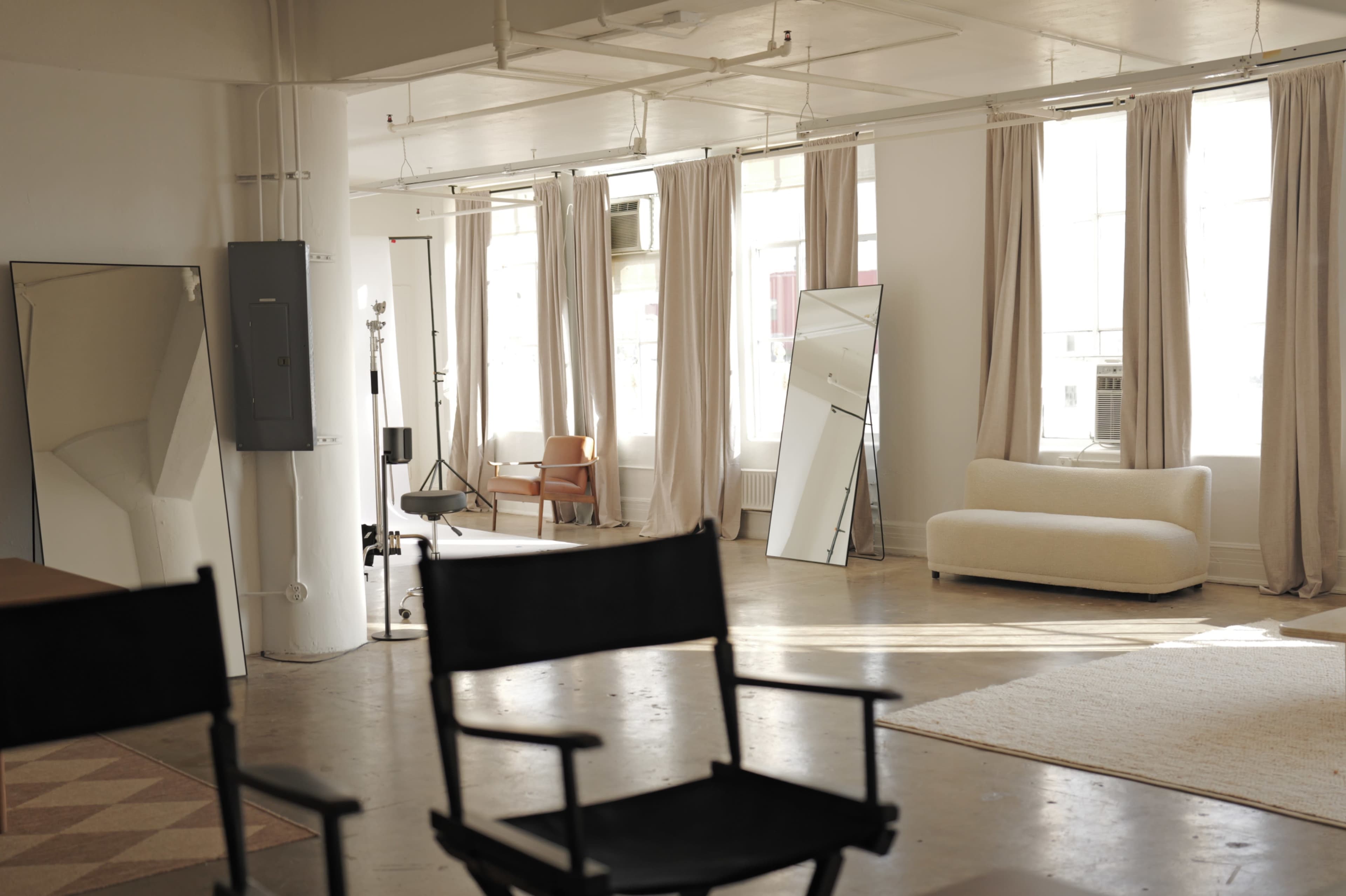 The image shows a bright, spacious interior with large windows, a mirror, a sofa, and a chair, all arranged around a light-colored rug.