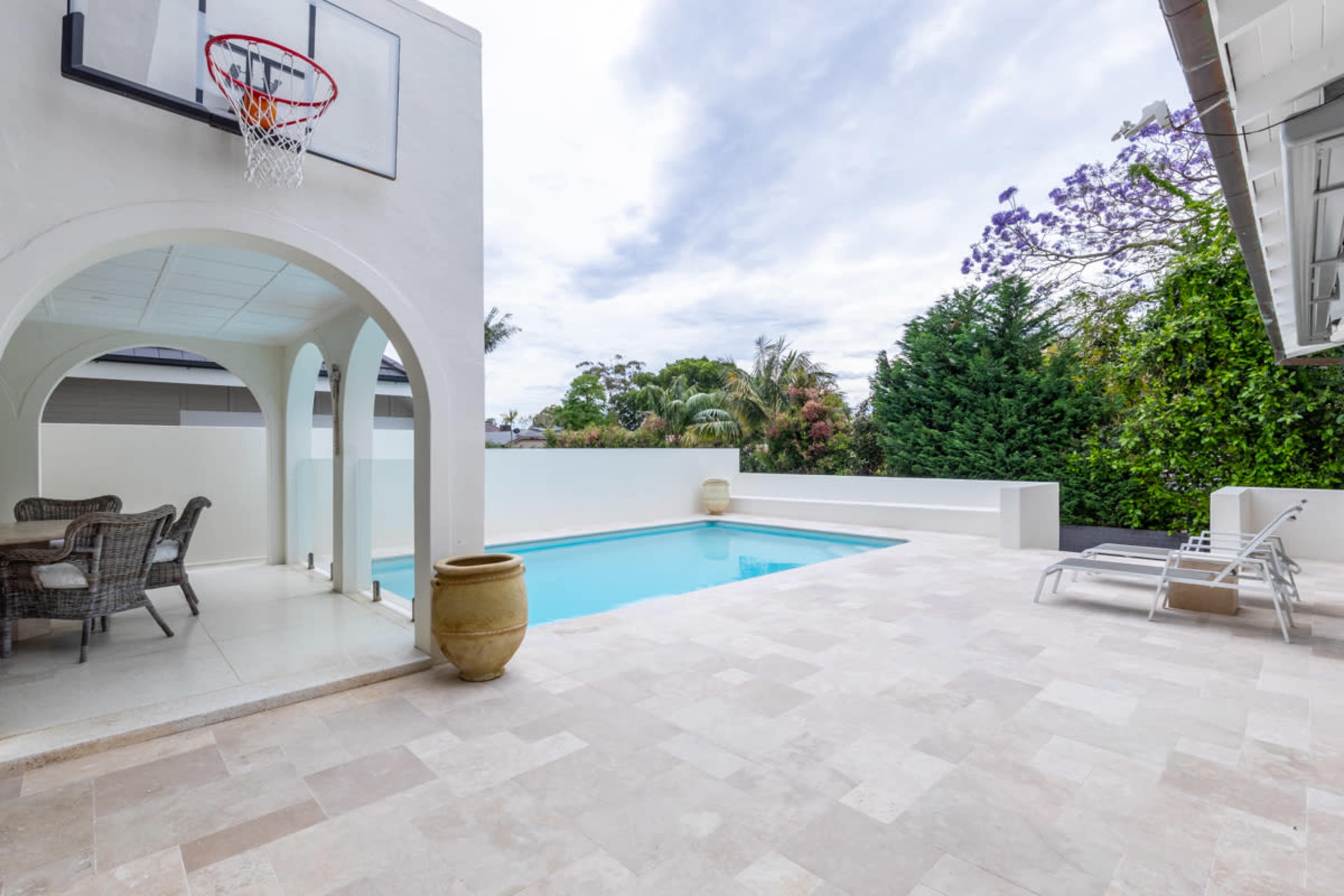A swimming pool surrounded by beige stone tiles features a basketball hoop and outdoor seating under an archway.
