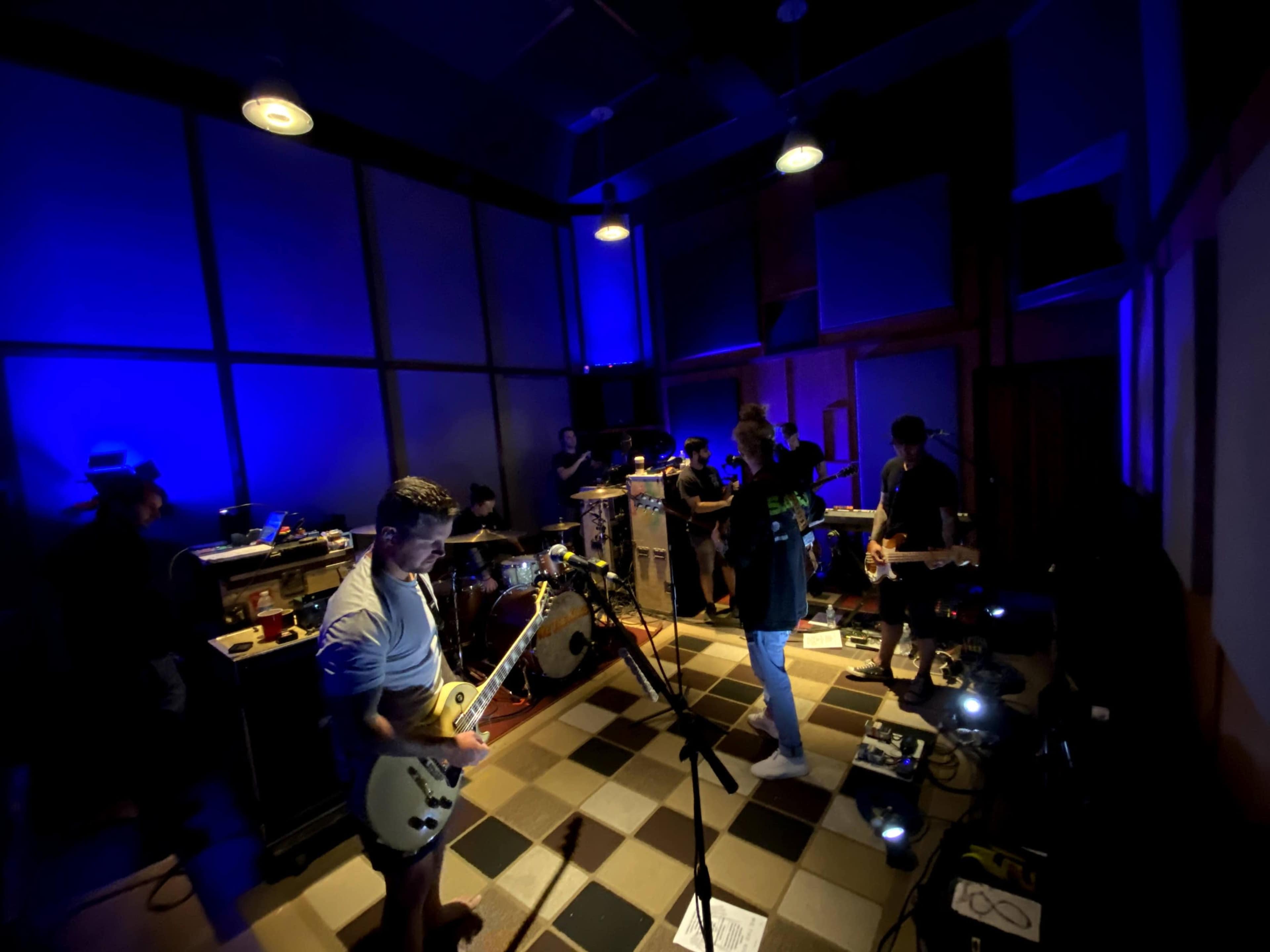 A group of musicians performs in a dimly lit studio, surrounded by instruments and amplifiers.