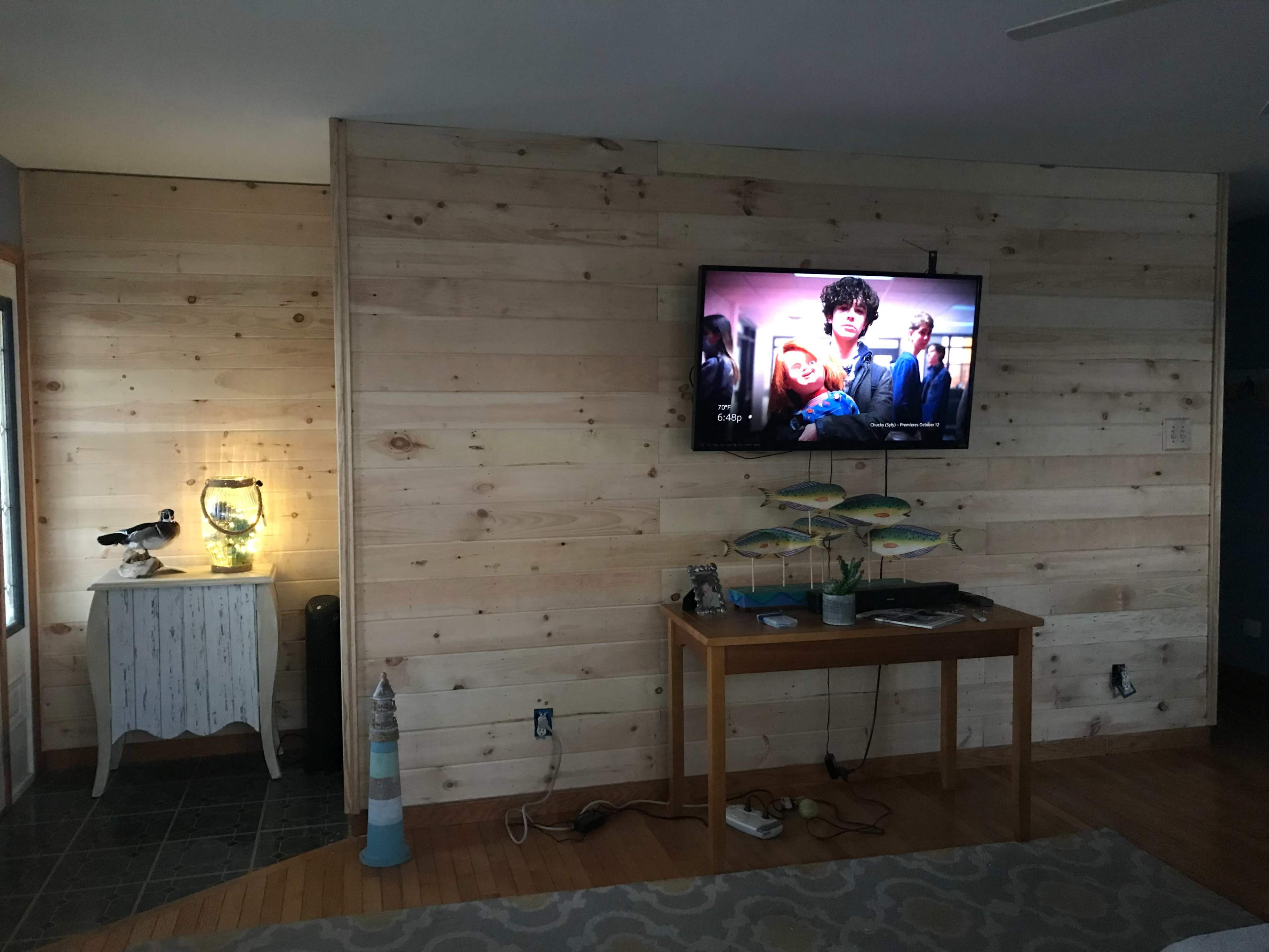 A wooden-paneled wall features a television displaying a scene from a movie, with a table and decorative items nearby.