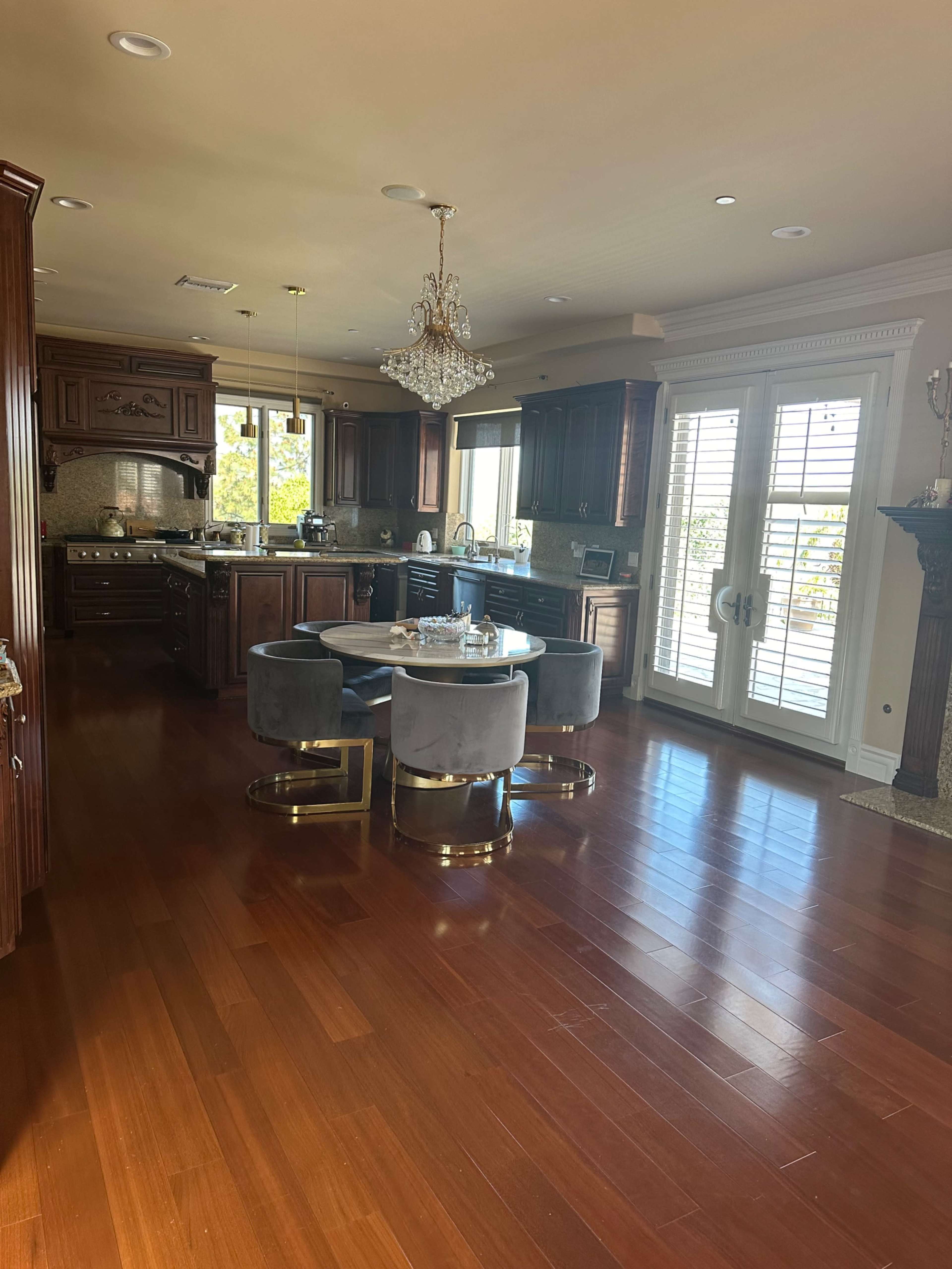 A spacious kitchen features dark wooden cabinets, a circular dining table with gray upholstered chairs, and large windows with shutters.