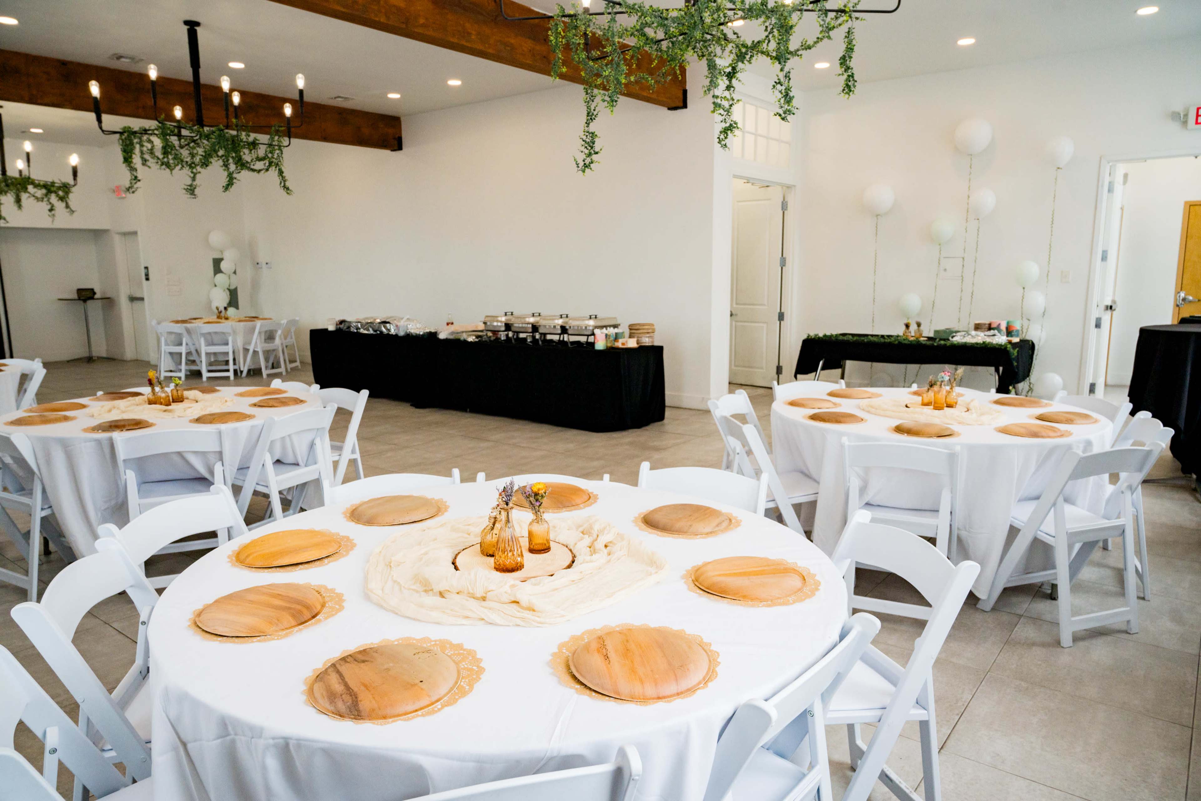 The image shows a spacious event room with several round tables set with white tablecloths and wooden chargers, alongside a buffet table covered with a black cloth in the background.